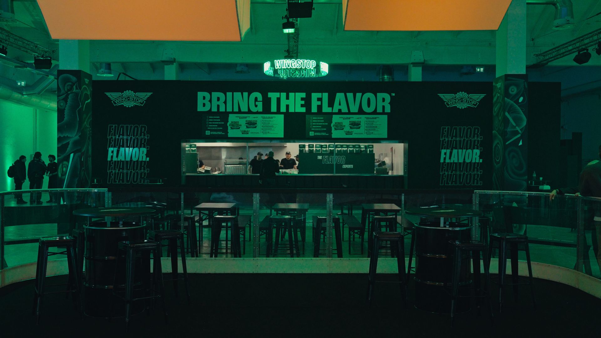 Indoor Wingstop counter under teal lighting, with a large "BRING THE FLAVOR" sign, menu boards above, staff at the prep area, and rows of stools and tables in the foreground.