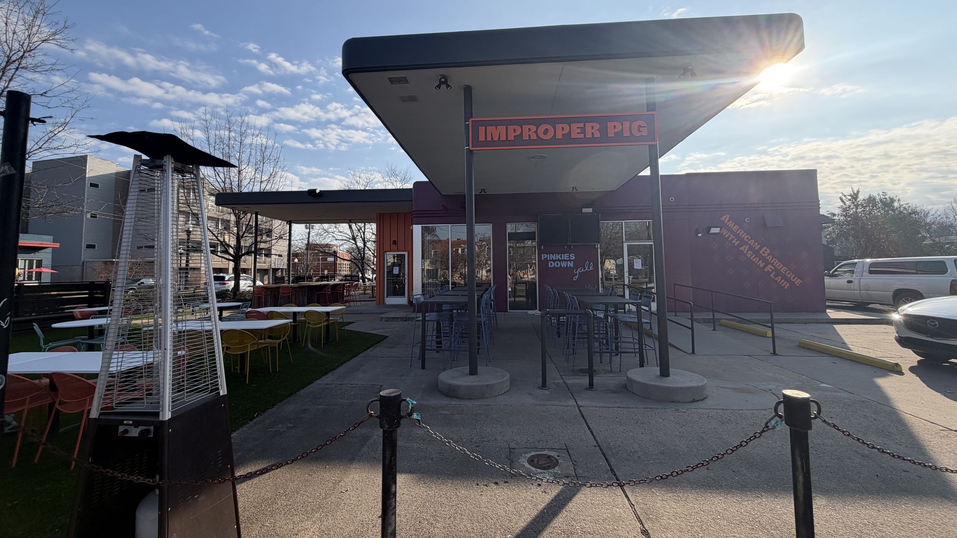 Outdoor seating area with colorful chairs and tables in front of a restaurant named "Improper Pig," featuring a sign that reads "American Barbecue with Asian Flair" under a sunny blue sky.