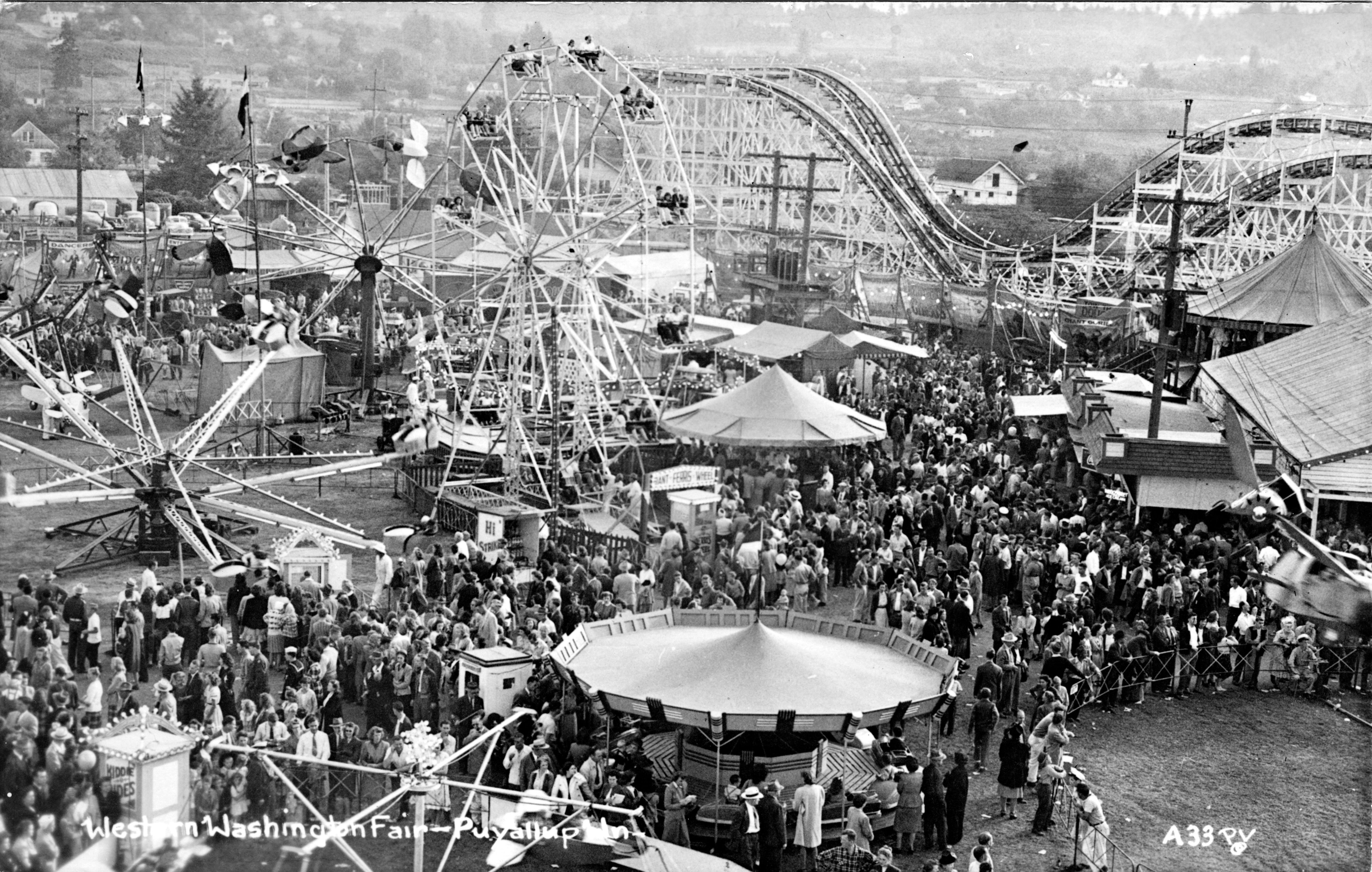 A black and white image featuring a crowd around rides including a ferris wheel and rollercoaster.