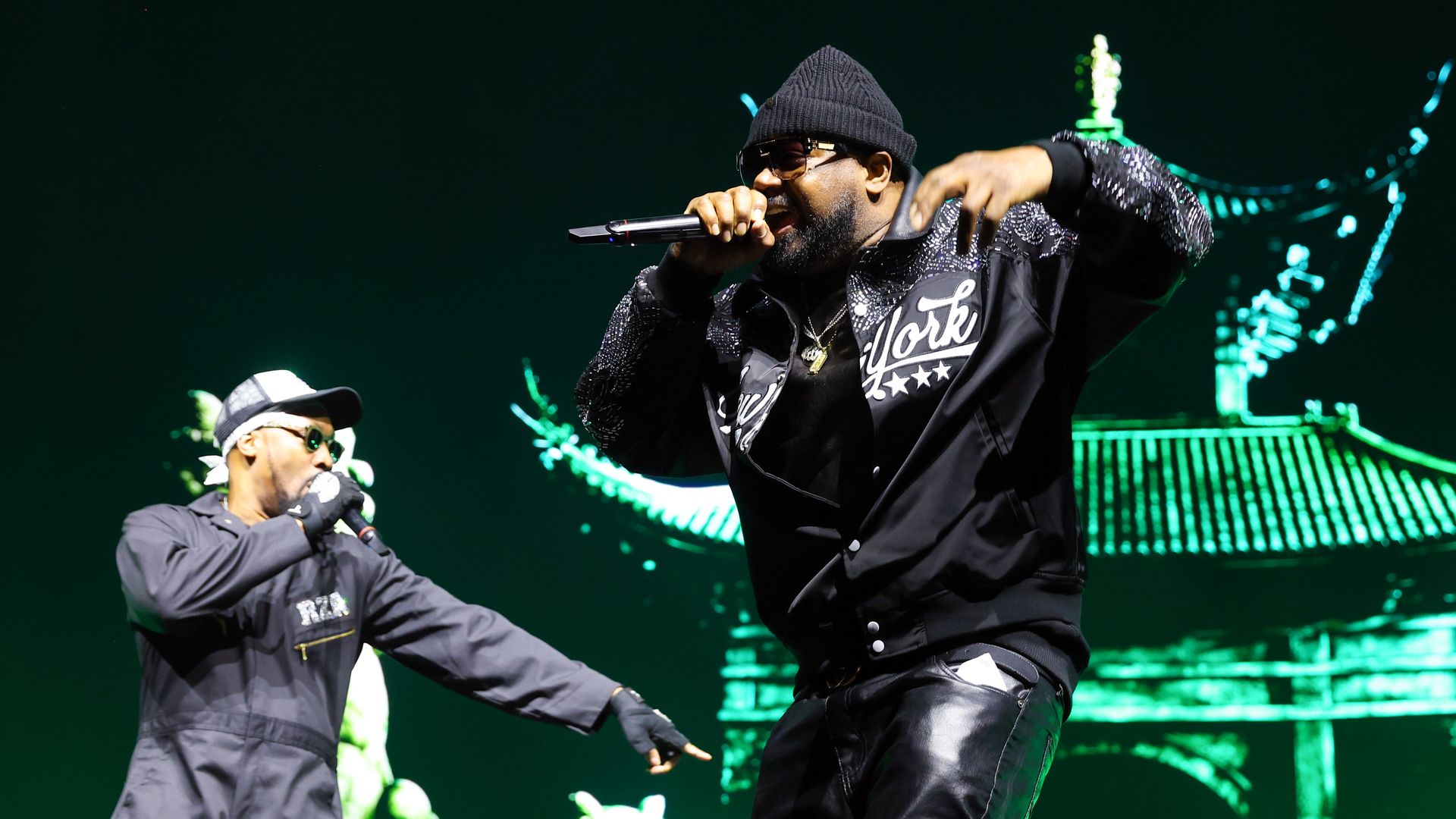 Two male hip-hop performers on a neon green stage; center rapper wears a black beanie and sunglasses, rapping into a microphone, while the other in a gray outfit and cap gestures toward the crowd.