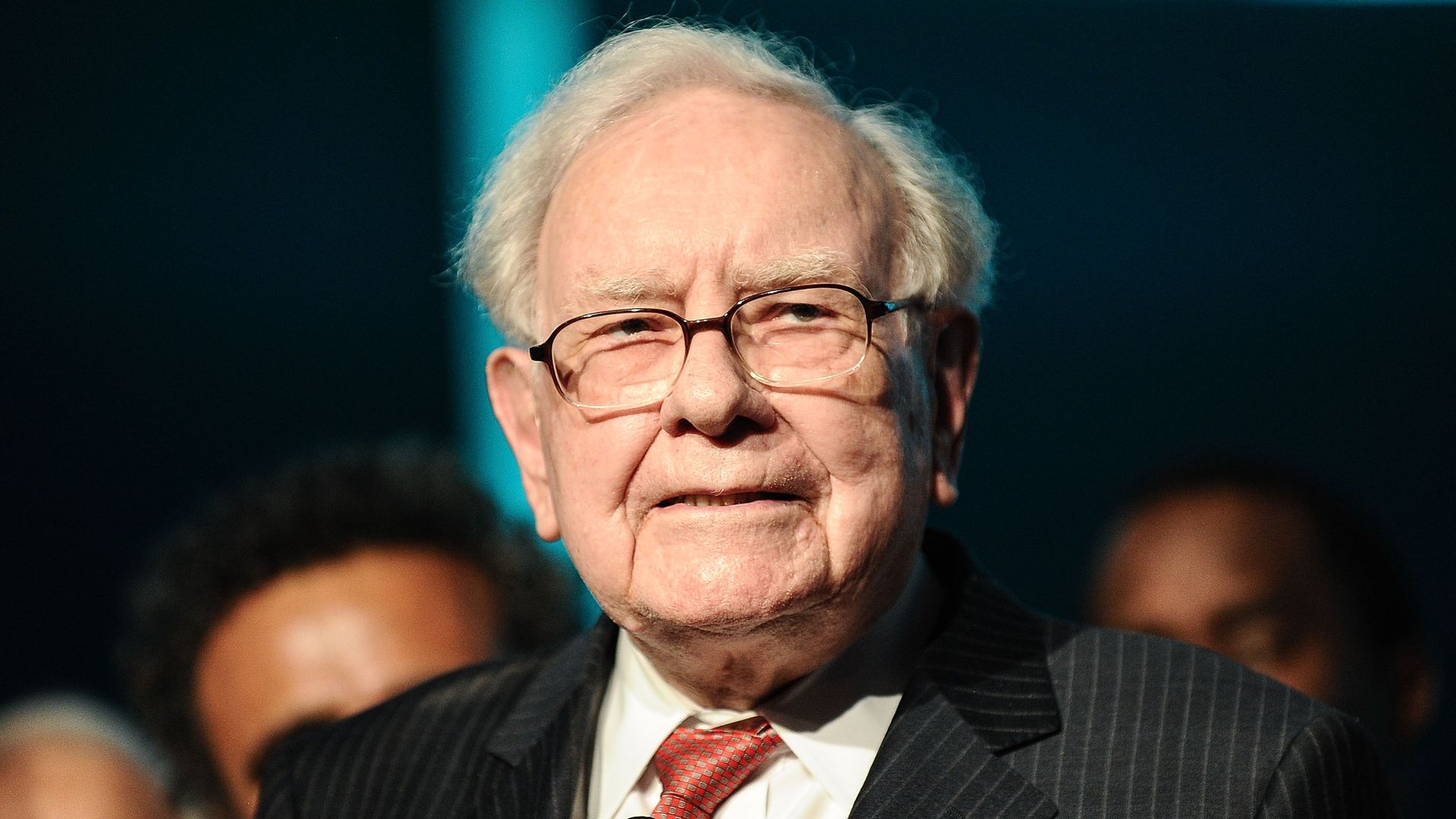 In this image, Warren Buffet stands in a suit and tie and looks at the camera.