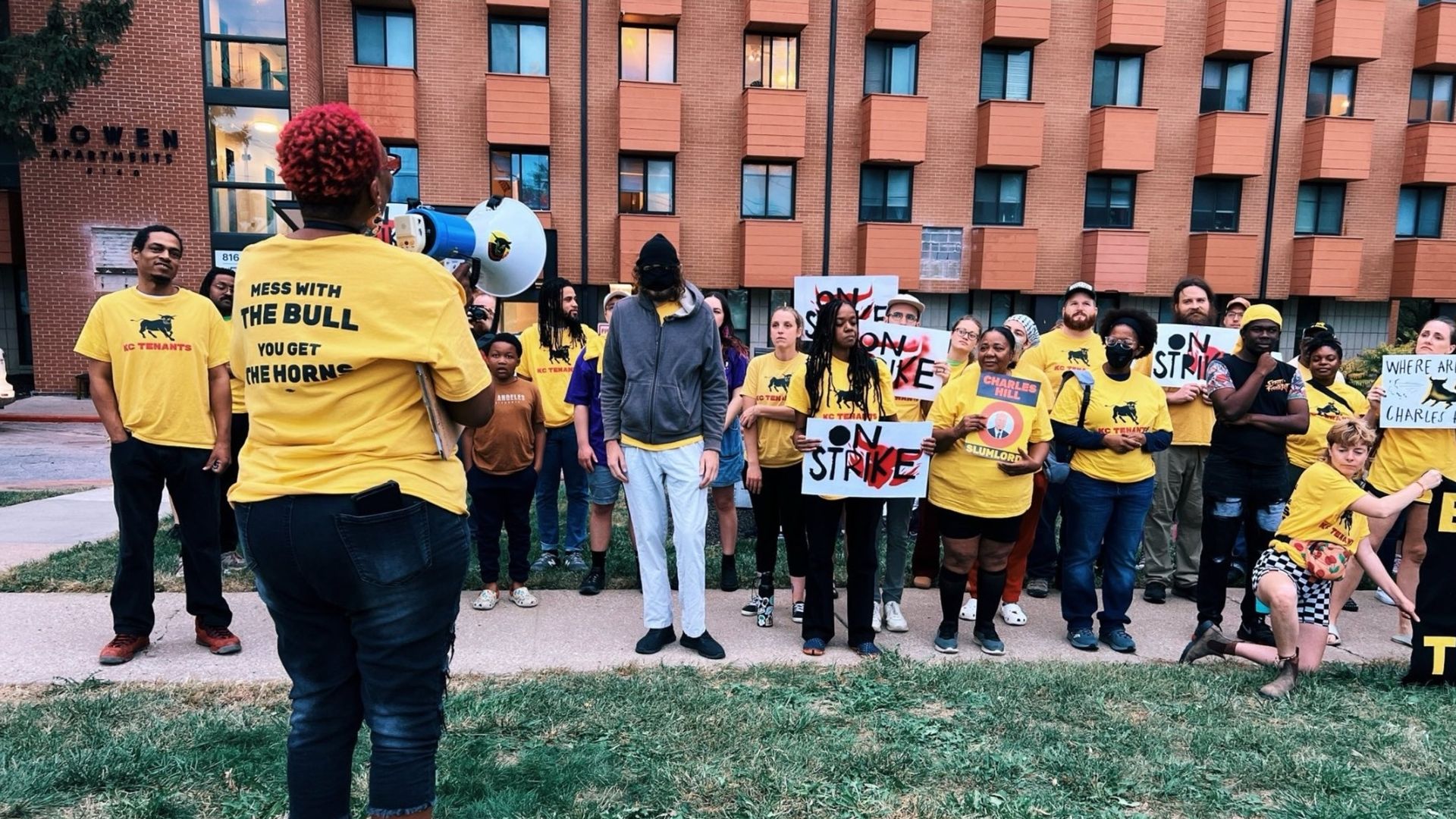 Group of protesters wearing yellow KC Tenants shirts, some holding signs reading "ON STRIKE" and "CHARLES HILL SLUMLORD," standing in front of a brick apartment building with a sign saying "Bowen Apartments."