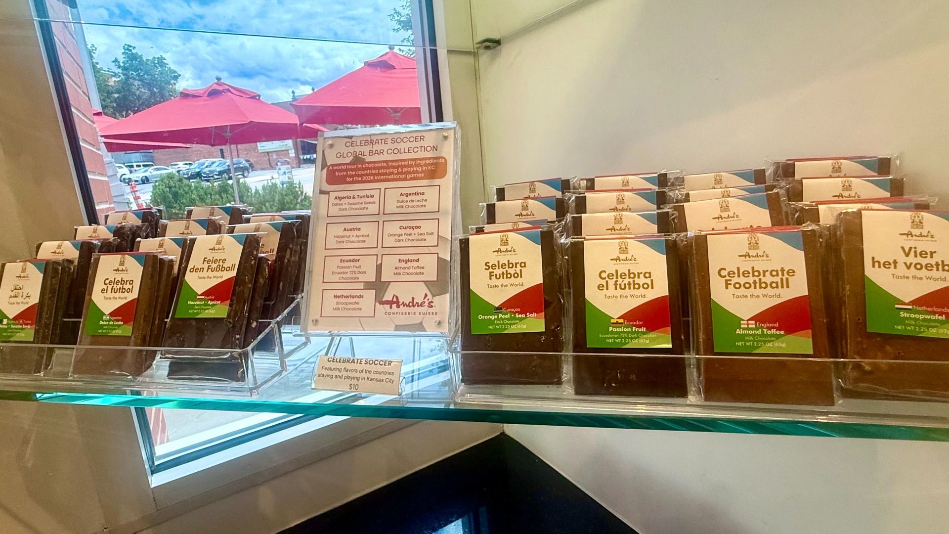 Display case of chocolate bars with white wrappers, blue and red accents; labels read "Celebrate Football" and "Celebra el fútbol". A flavor board and red outdoor umbrellas are visible outside.