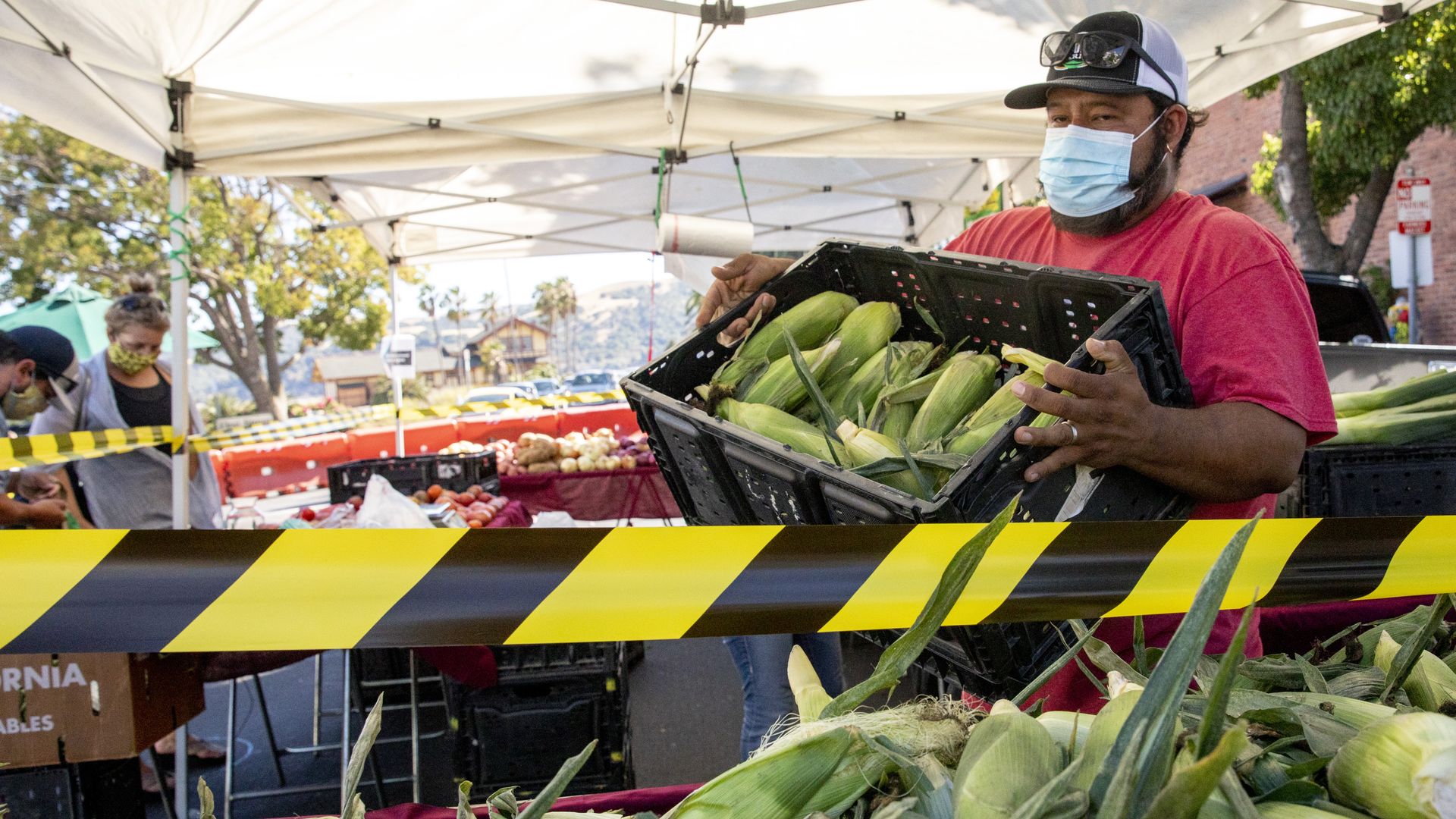 A man wearing a mask carries a plastic container of corn 