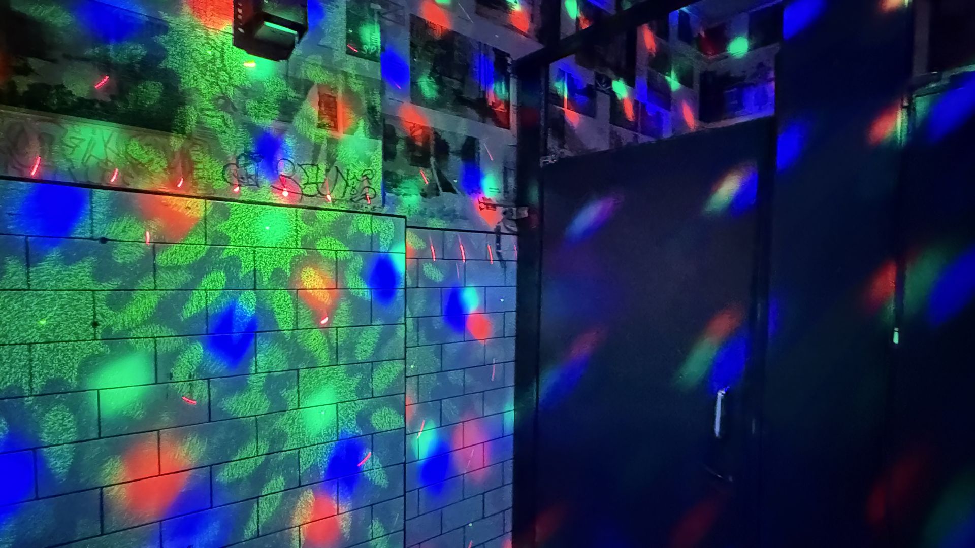 Dim room with a gray tiled wall covered in graffiti, illuminated by multicolor disco lights. Green, blue, and red reflections scatter across the tiles; a dark doorway on the right.