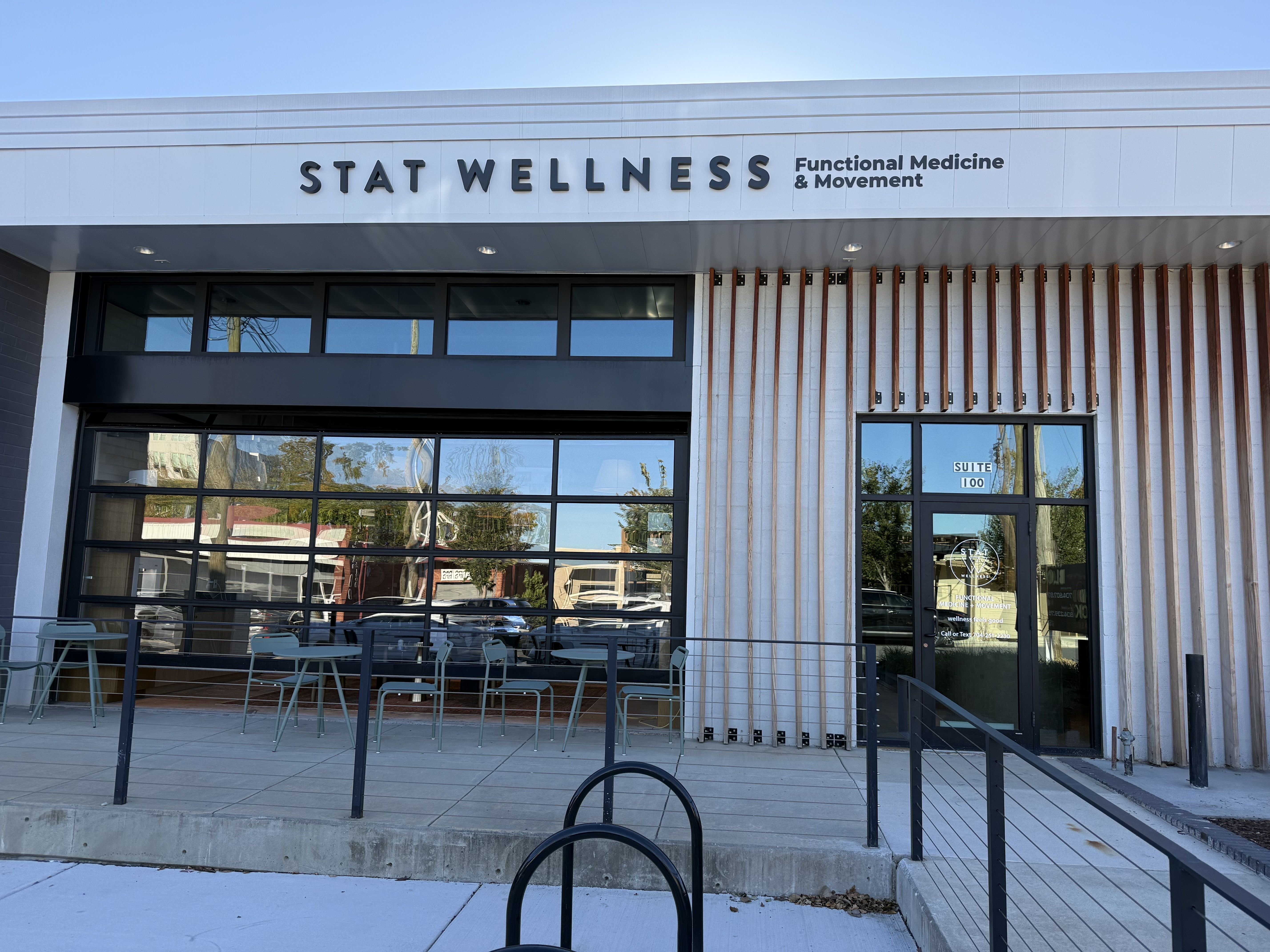 Front view of a modern wellness clinic with a white facade reading "STAT WELLNESS" and "Functional Medicine & Movement" on the sign, large glass doors, a wooden slat wall on the right, and outdoor seating.