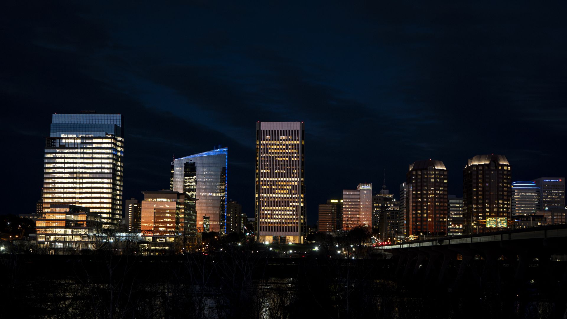 Night city skyline with tall glass buildings glowing white and amber lights along a dark blue sky. A riverfront foreground and distant highway signs complete the urban scene.