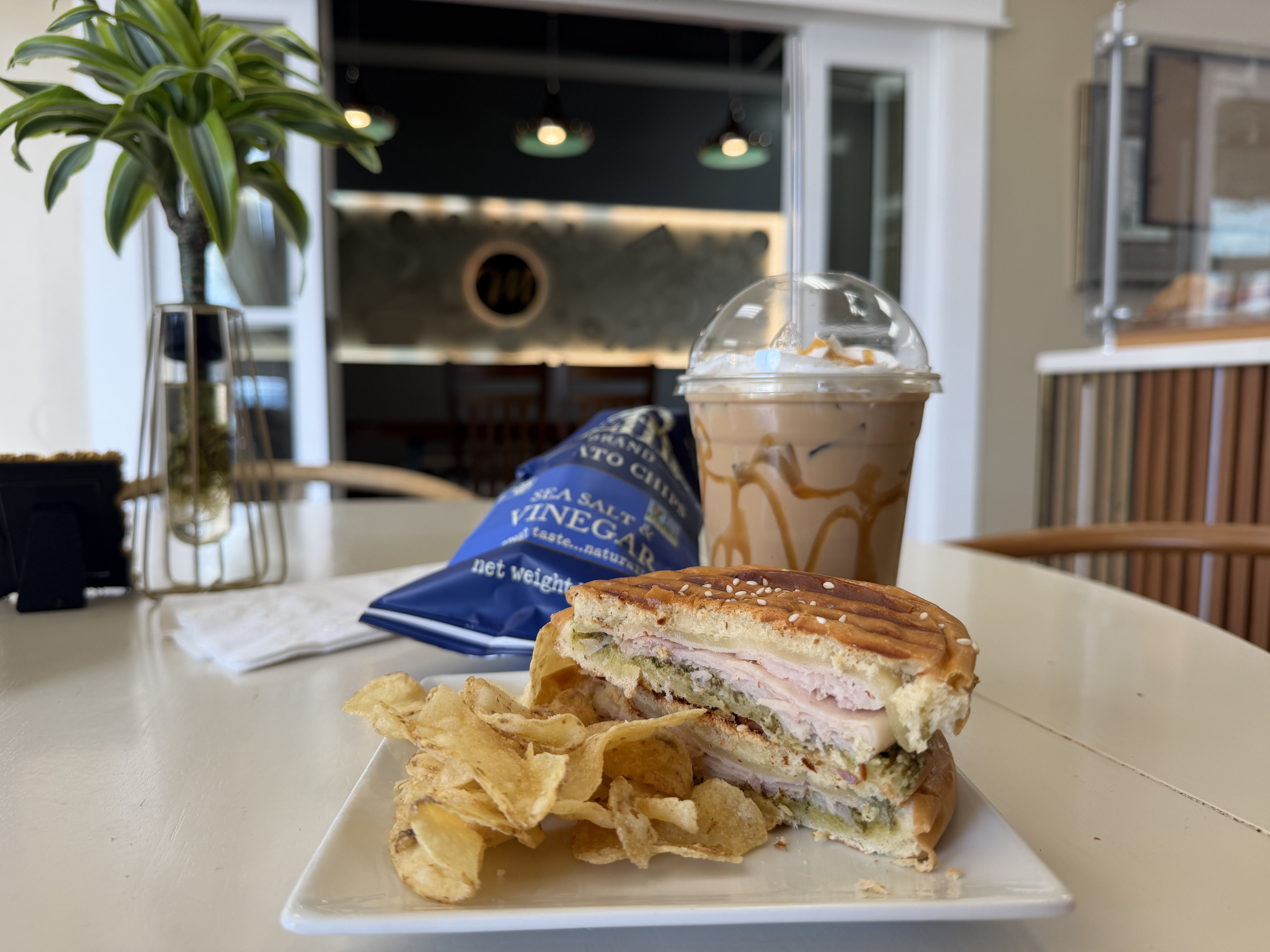 Sesame bun sandwich with turkey and cheese on a white plate, beside potato chips and a blue sea salt vinegar bag, plus an iced caramel coffee in a dome cup, in a bright cafe.