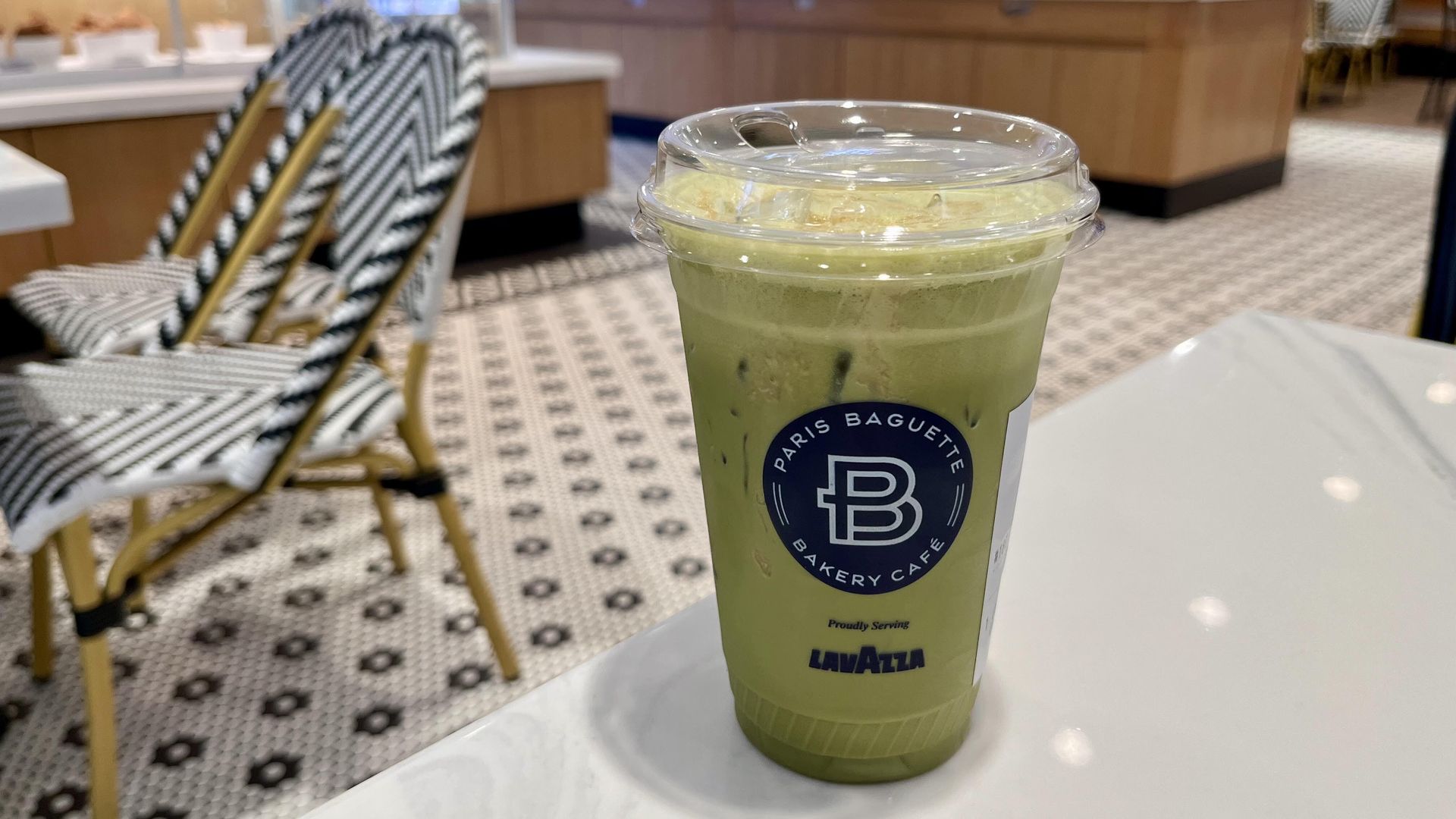 Green iced drink in a plastic cup with lid and Paris Baguette logo sits on a white table in a bakery cafe. Lavazza text visible; blurred display cases and chevron chairs in the background.