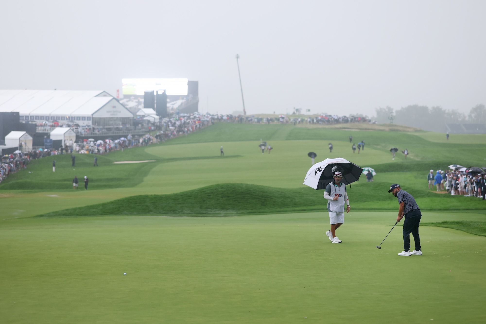 J.J. Spaun putts on the 18th green as it rains. His caddy walks behind him with an umbrella.