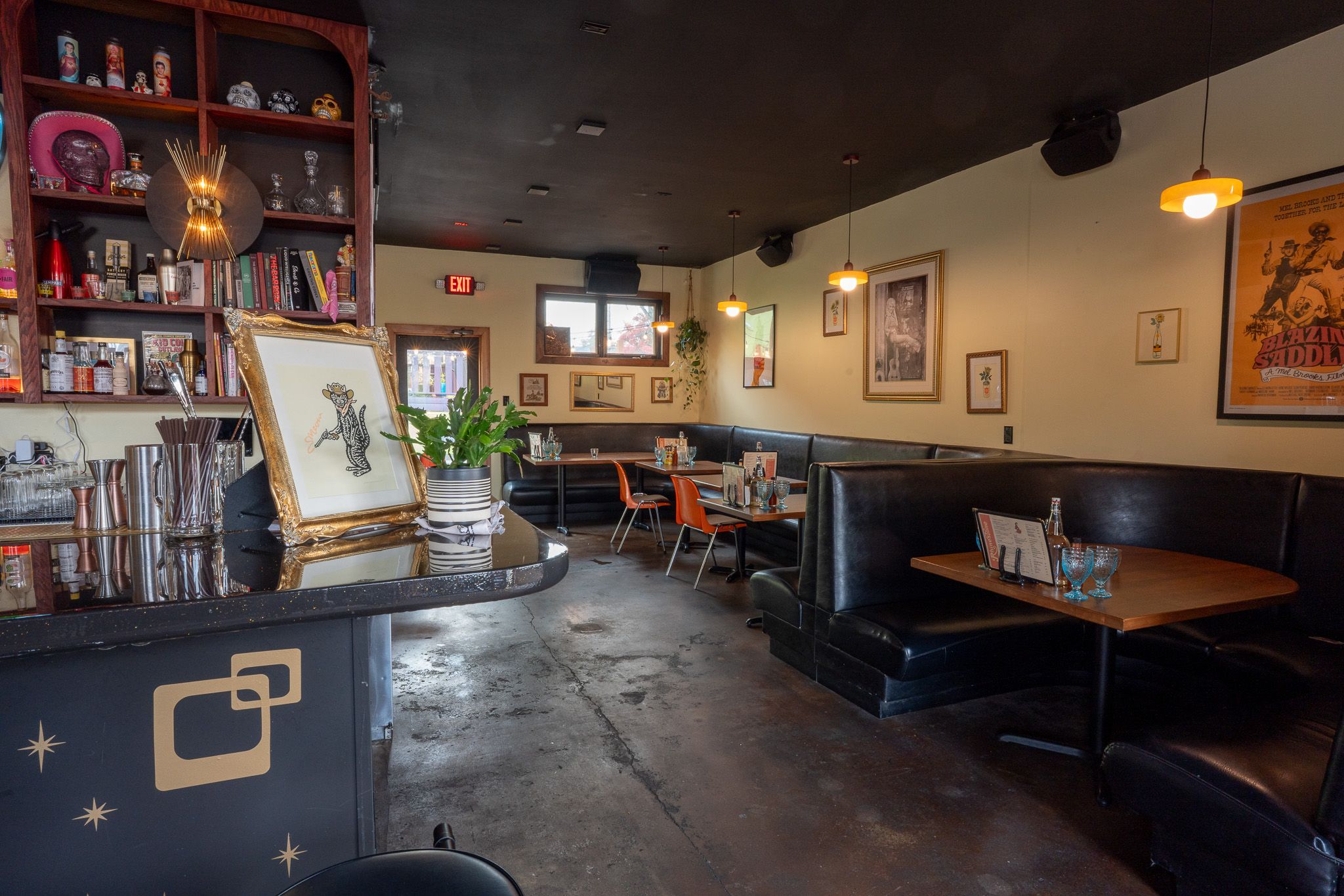 Cozy restaurant interior with black leather booths, wooden tables, orange chairs, hanging warm lights, framed art, a bar with shelves holding bottles and decor, and a plant on the counter.