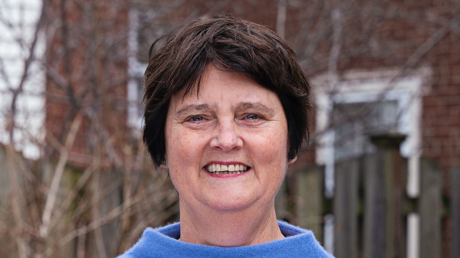 Smiling middle-aged woman with short dark hair wearing a light blue sweater outdoors, with a blurred background of leafless trees, wooden fence, and red brick building.