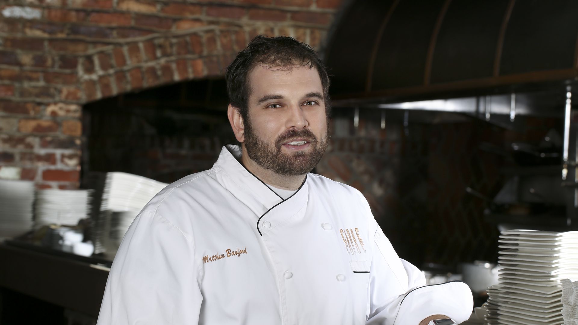 Bearded male chef in white coat with name "Matthew Basford" embroidered, standing in a kitchen with brick walls and stacks of white plates.