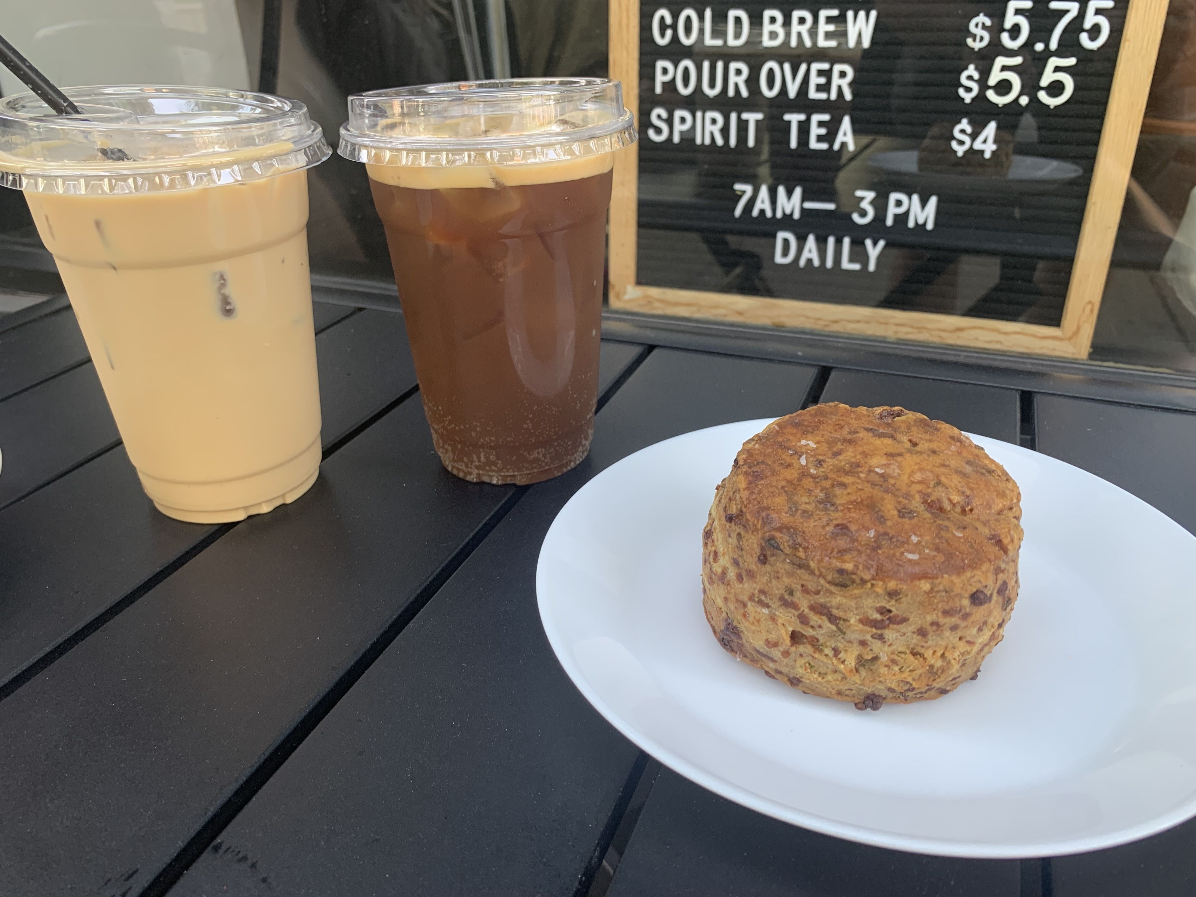 Two iced coffee drinks, one light and one dark, on a black table next to a white plate with a round baked pastry; a menu board shows prices for cold brew, pour over, and tea.