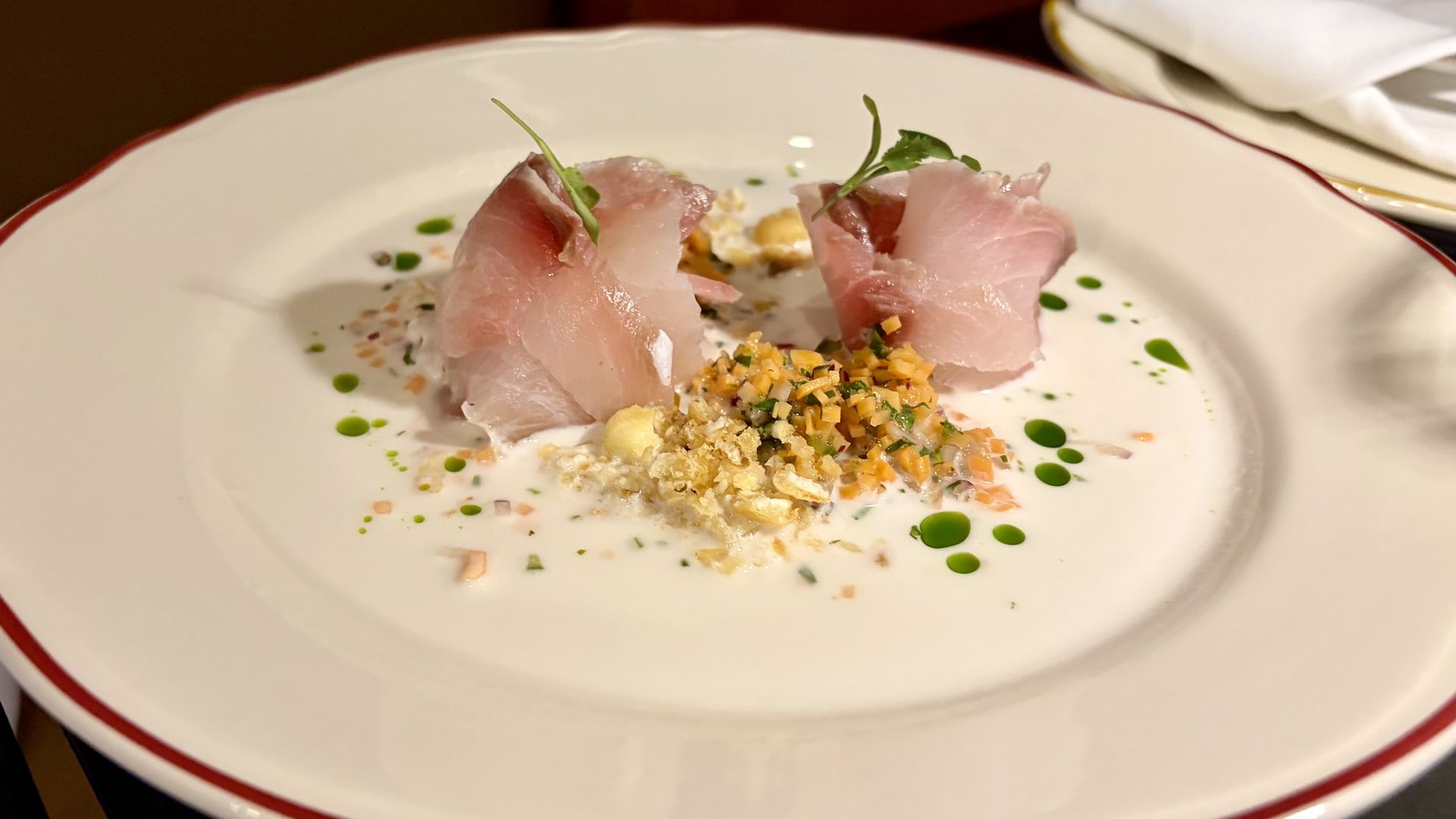 Elegant white plate with red rim holding a gourmet dish of two pink fish slices garnished with microgreens, surrounded by finely diced vegetables, crumbs, and green oil droplets in white sauce.