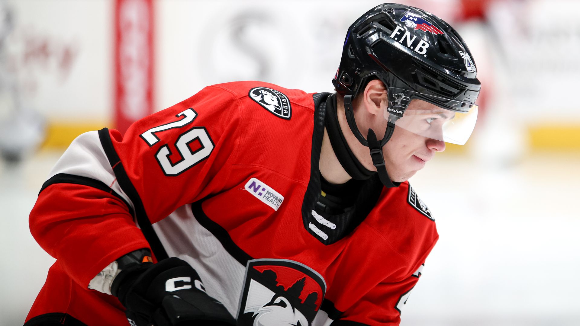 Hockey player in a red, white, and black uniform with number 29, wearing a black helmet with logos, focused on action during a game on ice.