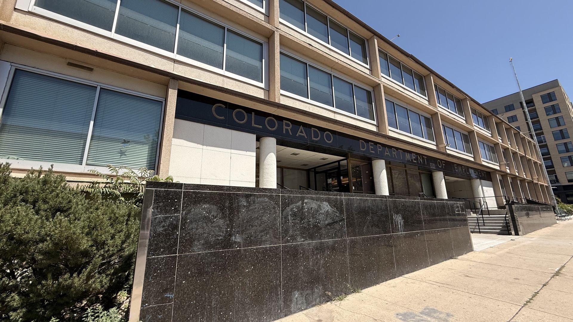 Exterior view of a beige multi-story office building with large glass windows, a dark marble base, and a sign for the Colorado Department of Employment above the entrance under a clear blue sky.