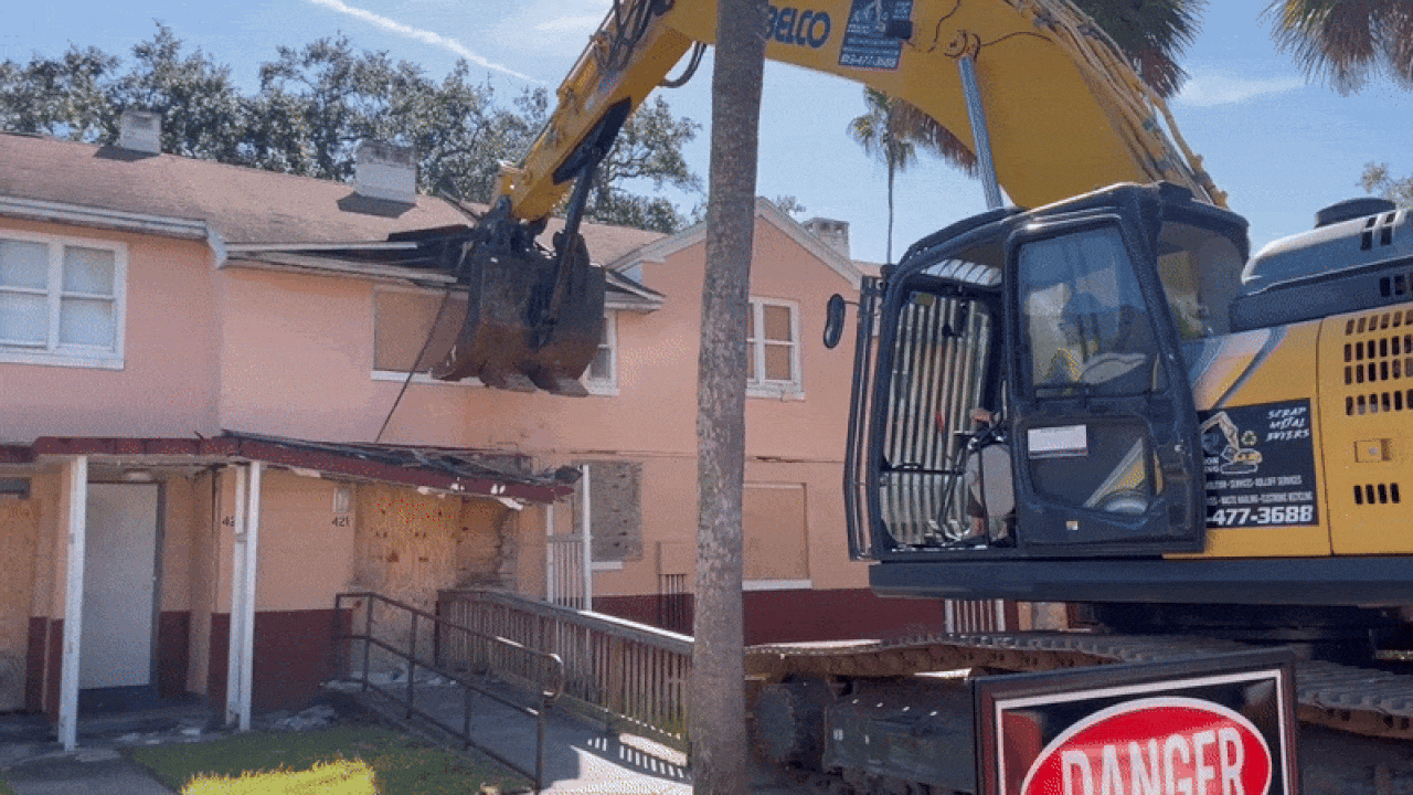 Yellow excavator demolishing roof of a peach-colored building with boarded windows, under clear blue sky and surrounded by palm trees. Danger sign visible on excavator base.