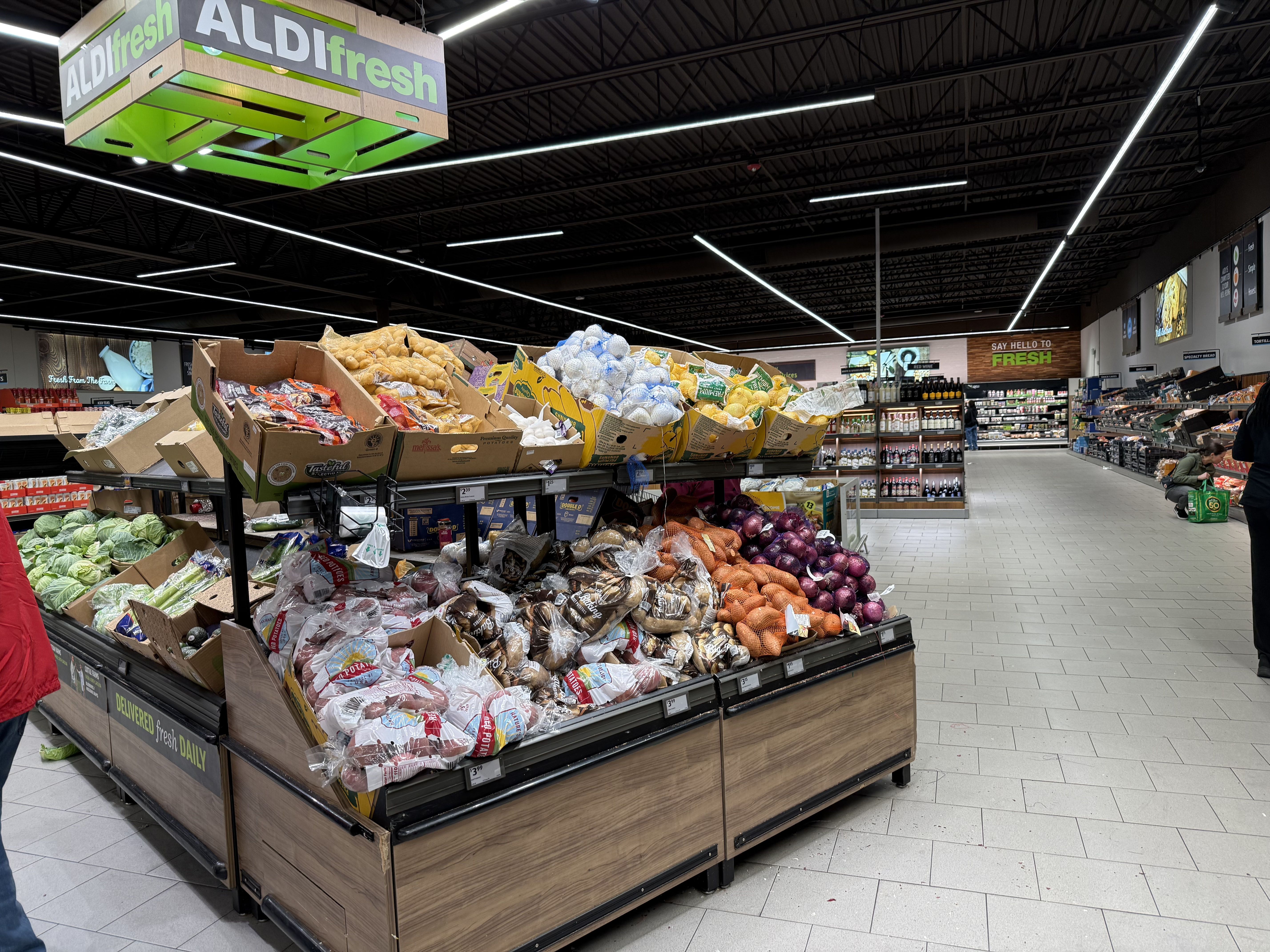 Produce section in an Aldi grocery store with various fresh vegetables and fruits displayed, including potatoes, onions, and cabbage under an "ALDI fresh" sign.