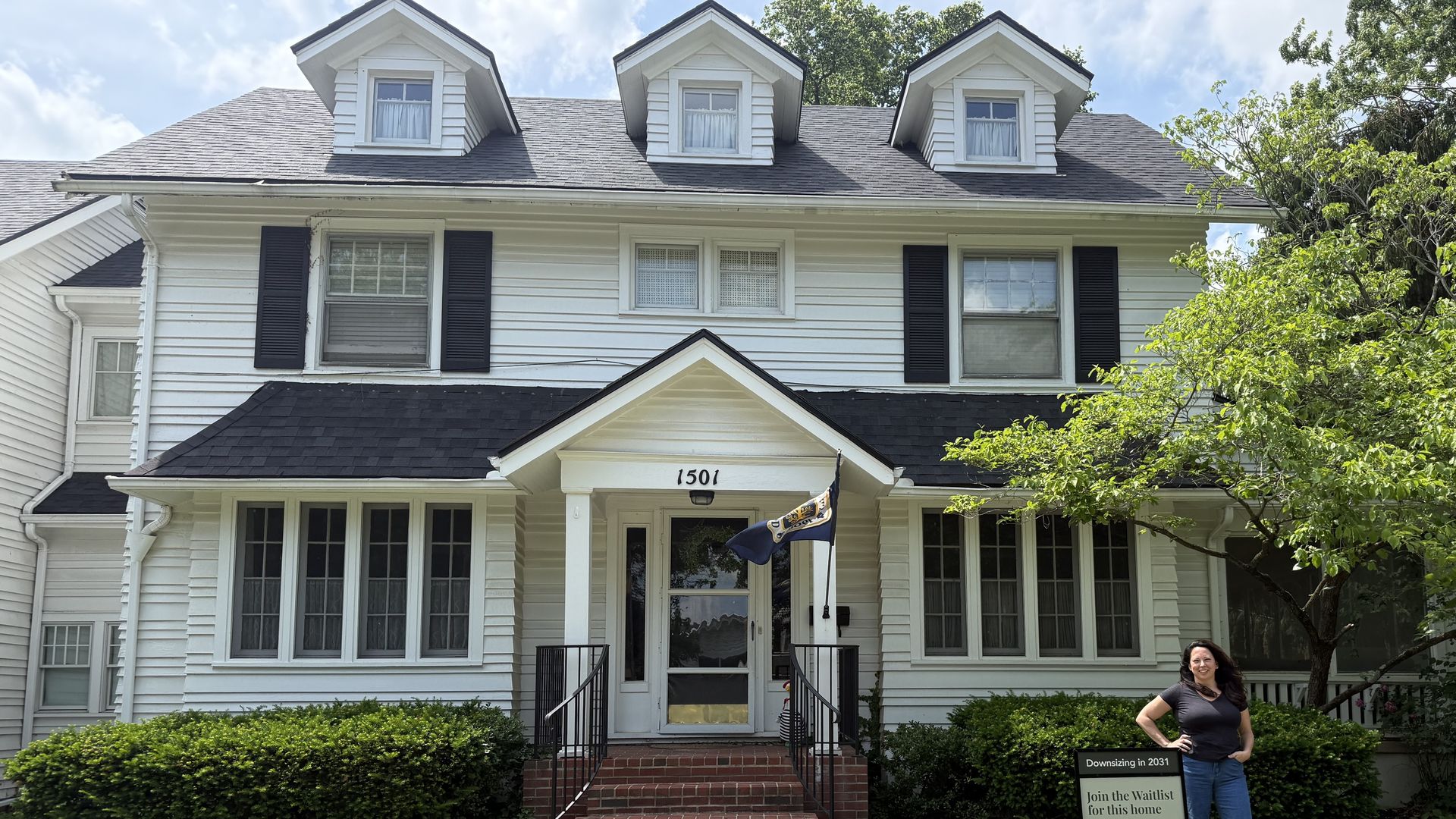 White two-story house with three dormer windows, black shutters, and a front porch. The address 1501 is above the door; a woman stands to the right beside a sign, with a tree and blue sky.