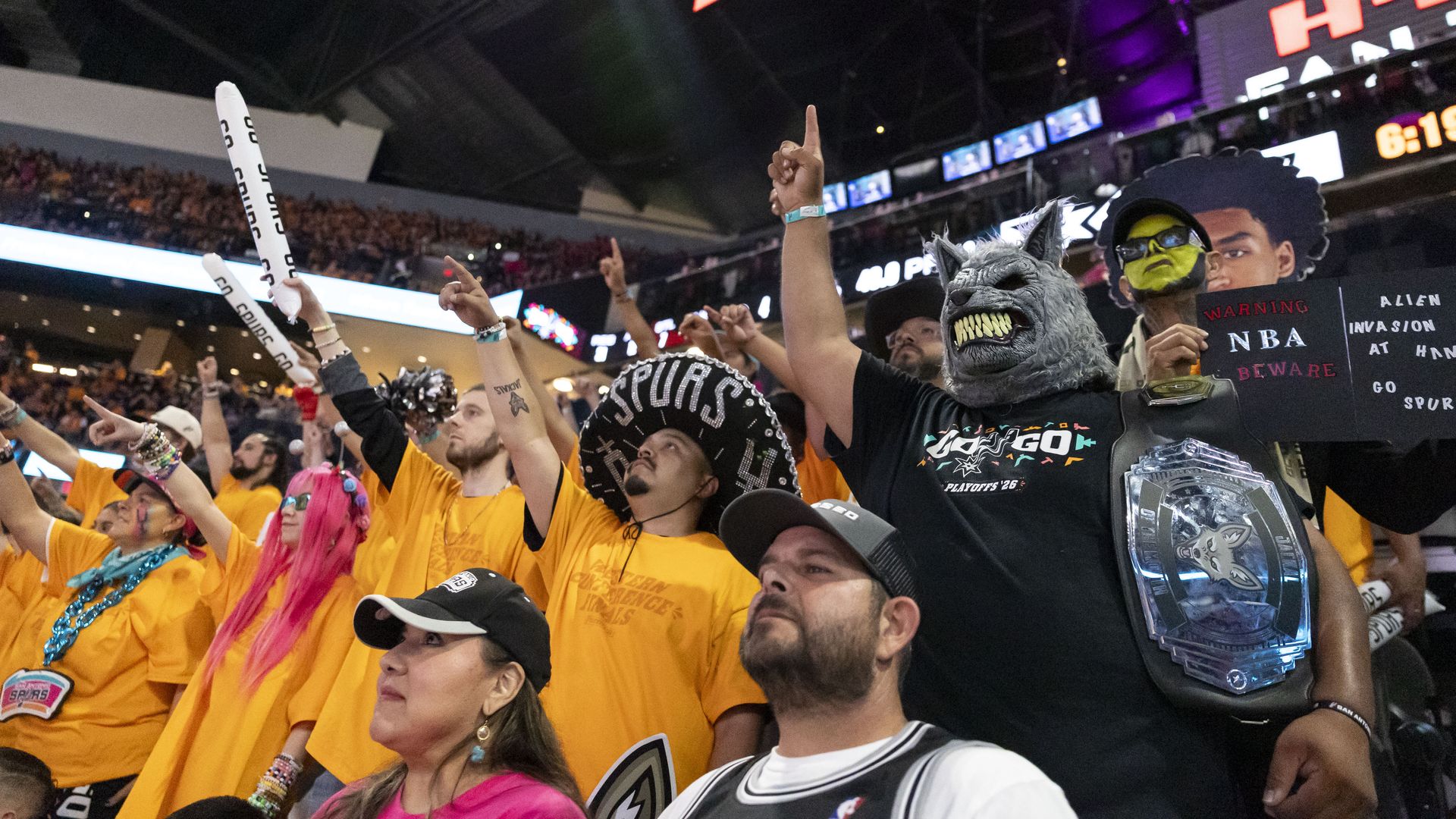 Packed arena crowd in yellow shirts cheers with foam sticks. A person wearing a gray wolf mask and a large championship belt stands front-right, with bright signs and screens in the background.