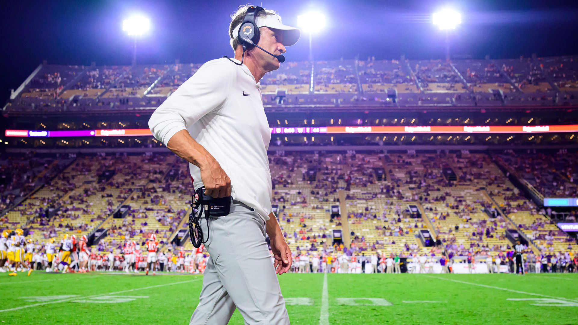Lane Kiffin, wearing a headset and visor, walks past the camera inside Tiger Stadium. The football field is behind him as players look ready to huddle up.