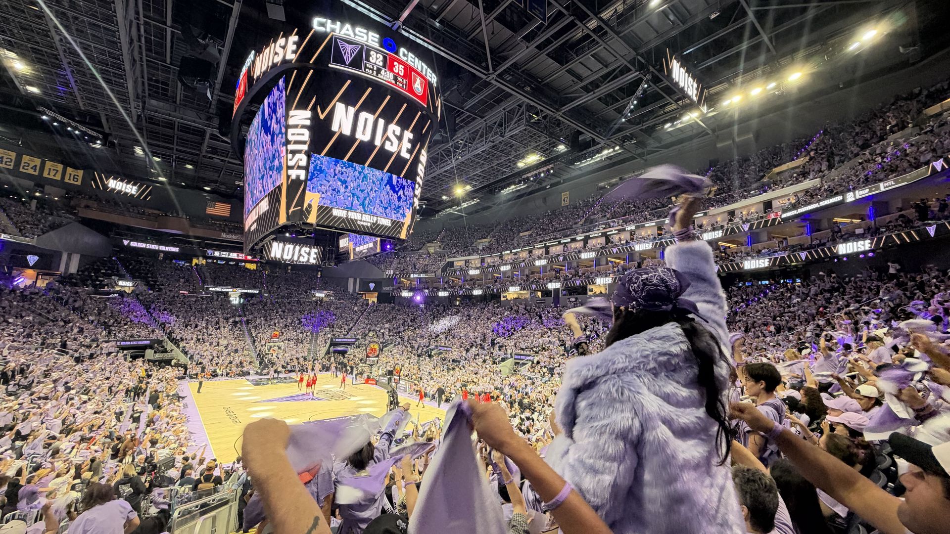 Inside a packed basketball arena at night, a large circular scoreboard hangs overhead displaying scores and the word NOISE. Fans in white fill the stands, with purple lighting and banners overhead.