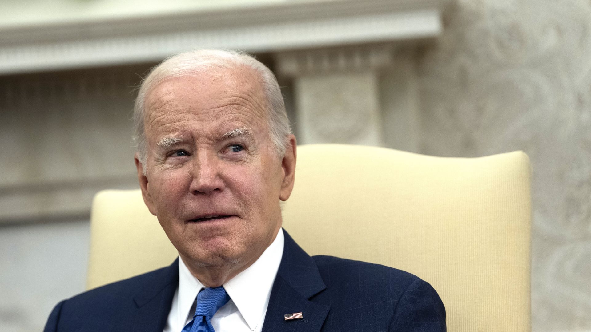 President Biden sits in a light yellow chair in the Oval Office.