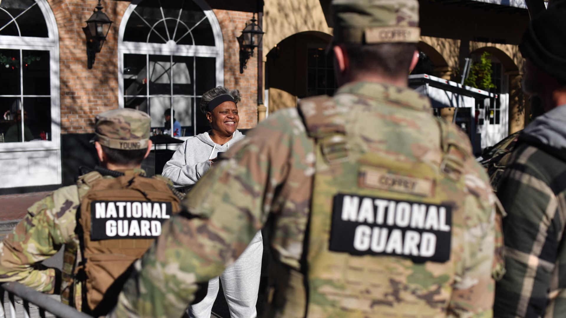 Two National Guard soldiers are viewed from behind as they talk with a woman in a grey sweatsuit.