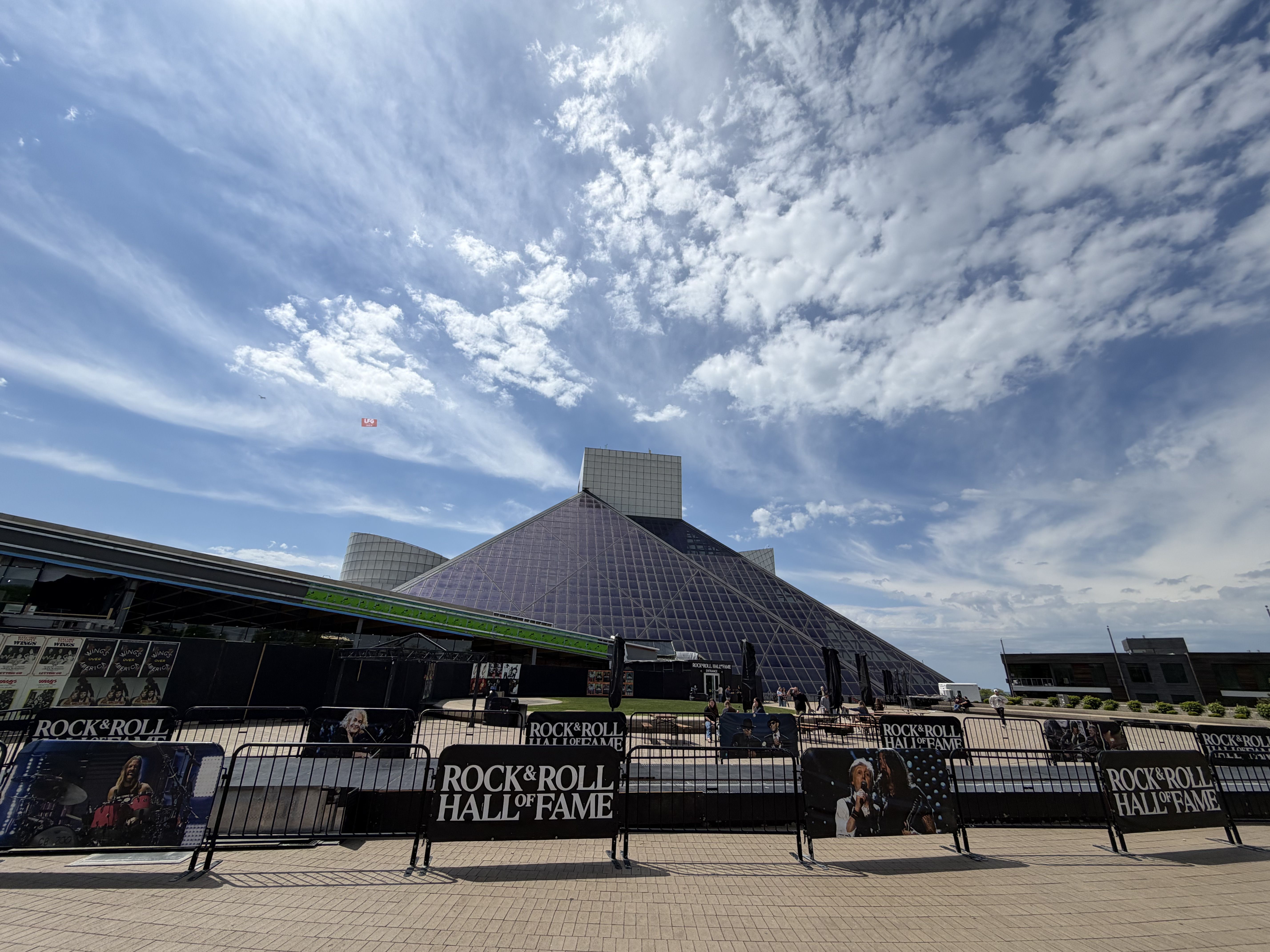 Bright blue sky with scattered white clouds above the Rock & Roll Hall of Fame glass pyramid. Foreground shows barriers and posters along a fenced plaza.