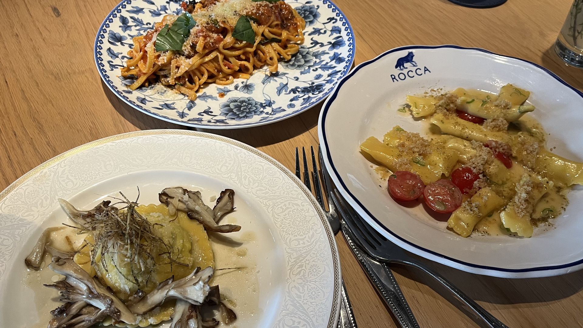Three dishes on a wooden table: a blue-and-white plate of tomato-basil spaghetti with parmesan, a Rocca-branded plate with creamy stuffed pasta and tomatoes, and a plate of mushrooms in a pale sauce.