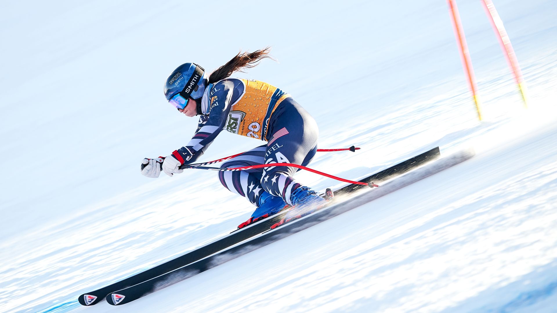 Female skier in blue USA suit and yellow bib racing downhill on snowy slope, wearing helmet and goggles, with ski poles angled behind her, passing orange gates.