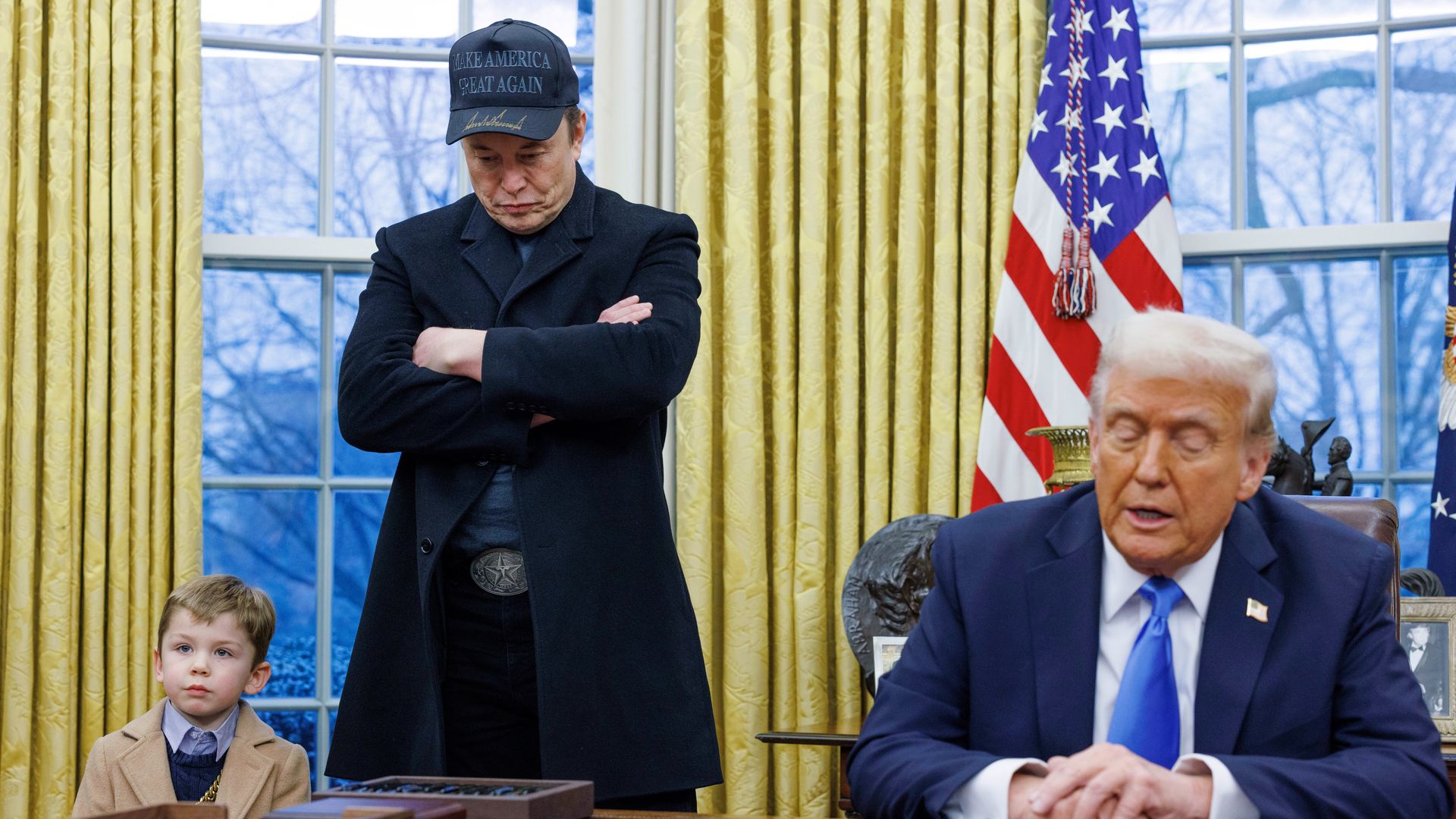 Elon Musk, wearing a black coat and black Make America Great Again, with his son standing next to him in a beige coat and Donald Trump seated behind the resolute desk wearing a blue suit.