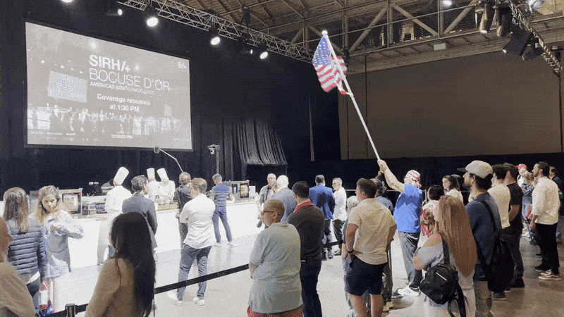 A man wearing a blue shirt waves a giant American flag as chefs cook in the background.