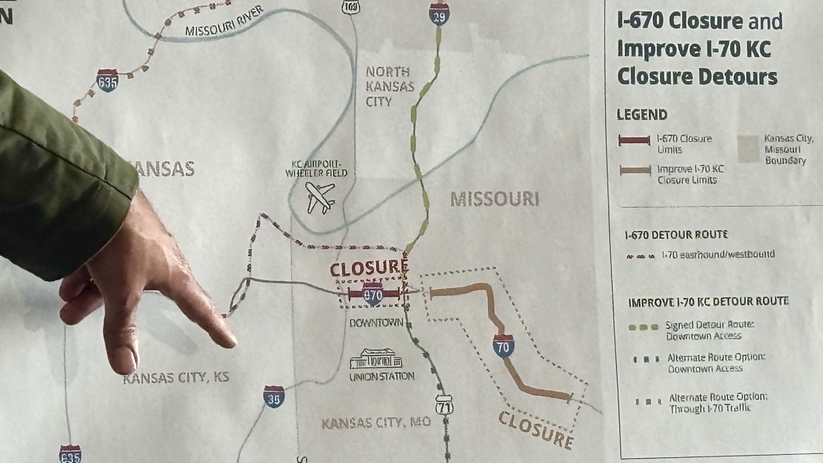 Map of the Kansas City area showing I-670 closure and I-70 detour routes; a hand points to downtown Kansas City near the airport, Missouri River, and North Kansas City.