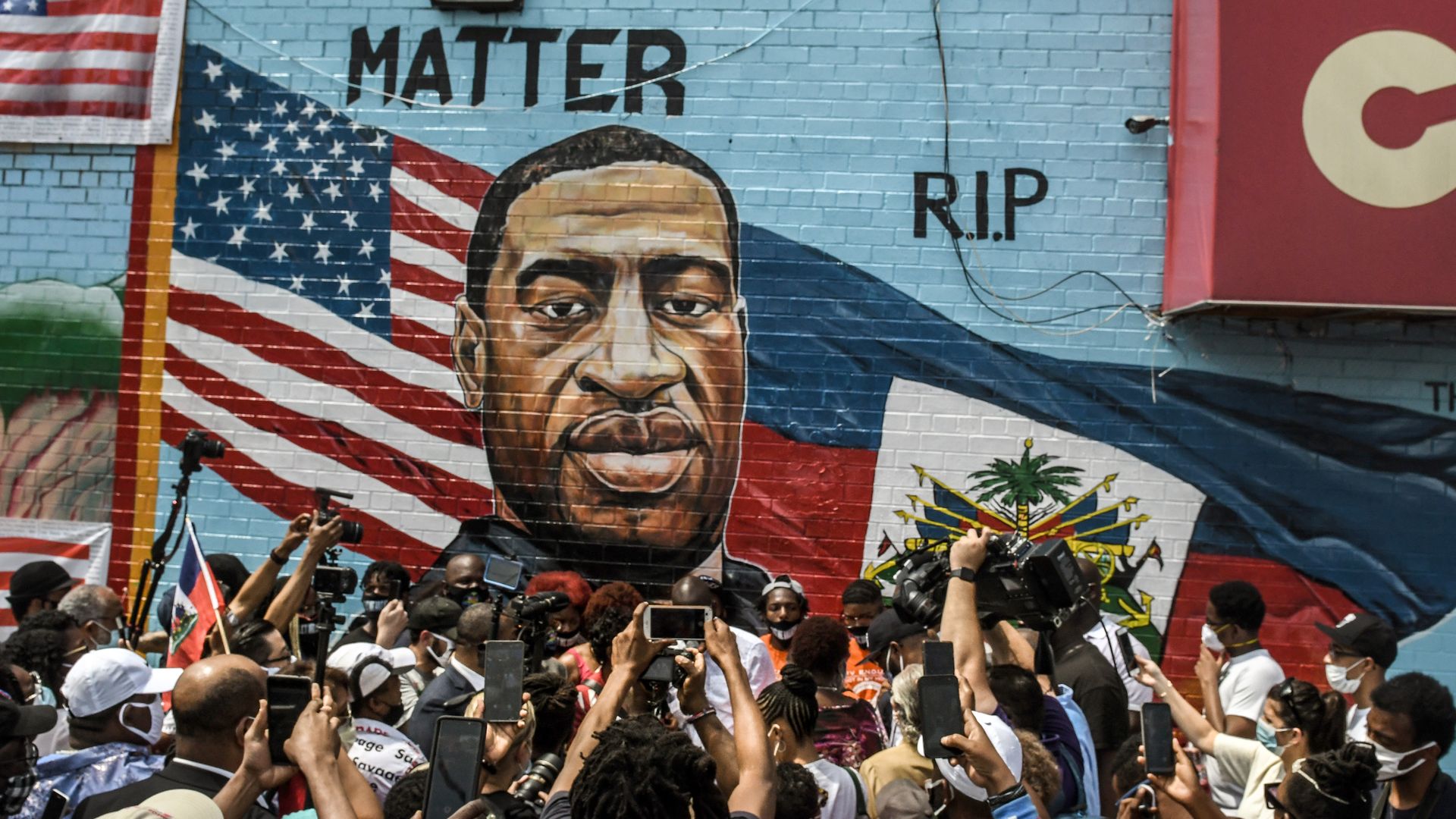 Dozens stand in front of a mural for George Floyd