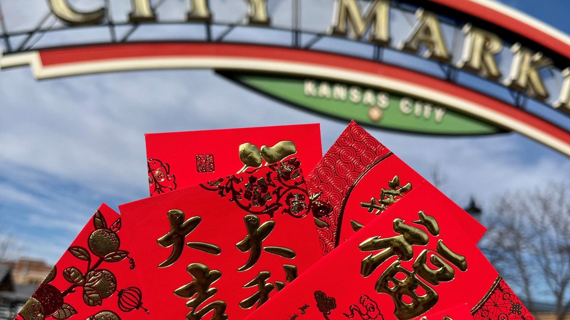 Close-up of red envelopes with gold Chinese characters and designs held up against a blurred background arch that says "City Market Kansas City" under a partly cloudy blue sky.
