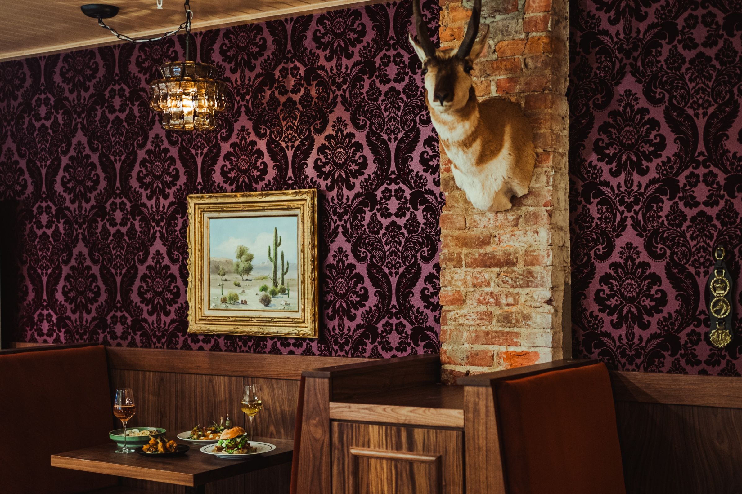 Cozy dining area with dark purple damask wallpaper, a brick column, and a mounted llama head. A gold-framed desert painting hangs beside a glass lantern; tables display plates of appetizers and wine.