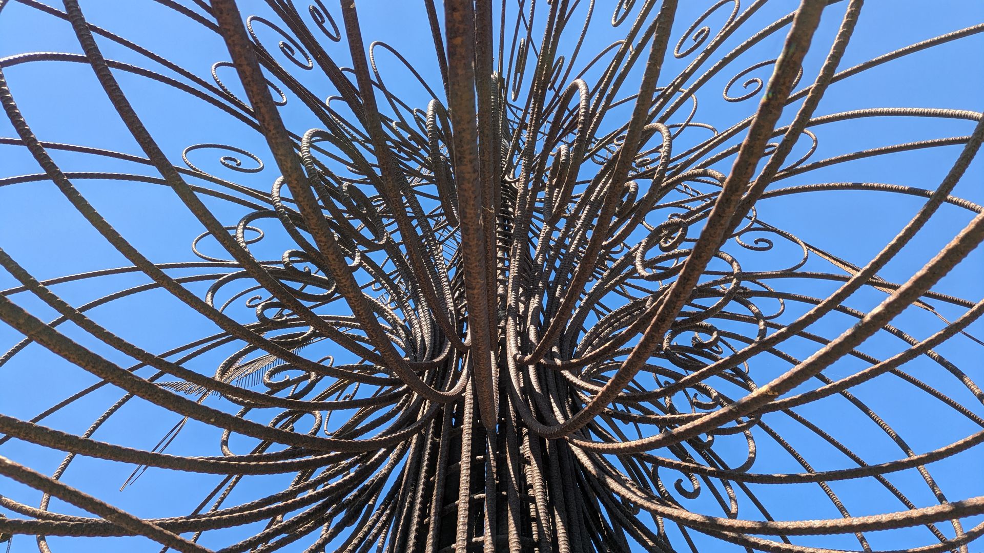 Close-up of a rusted metal sculpture with numerous curved, spiraling bars radiating from a central core, set against a clear blue sky.