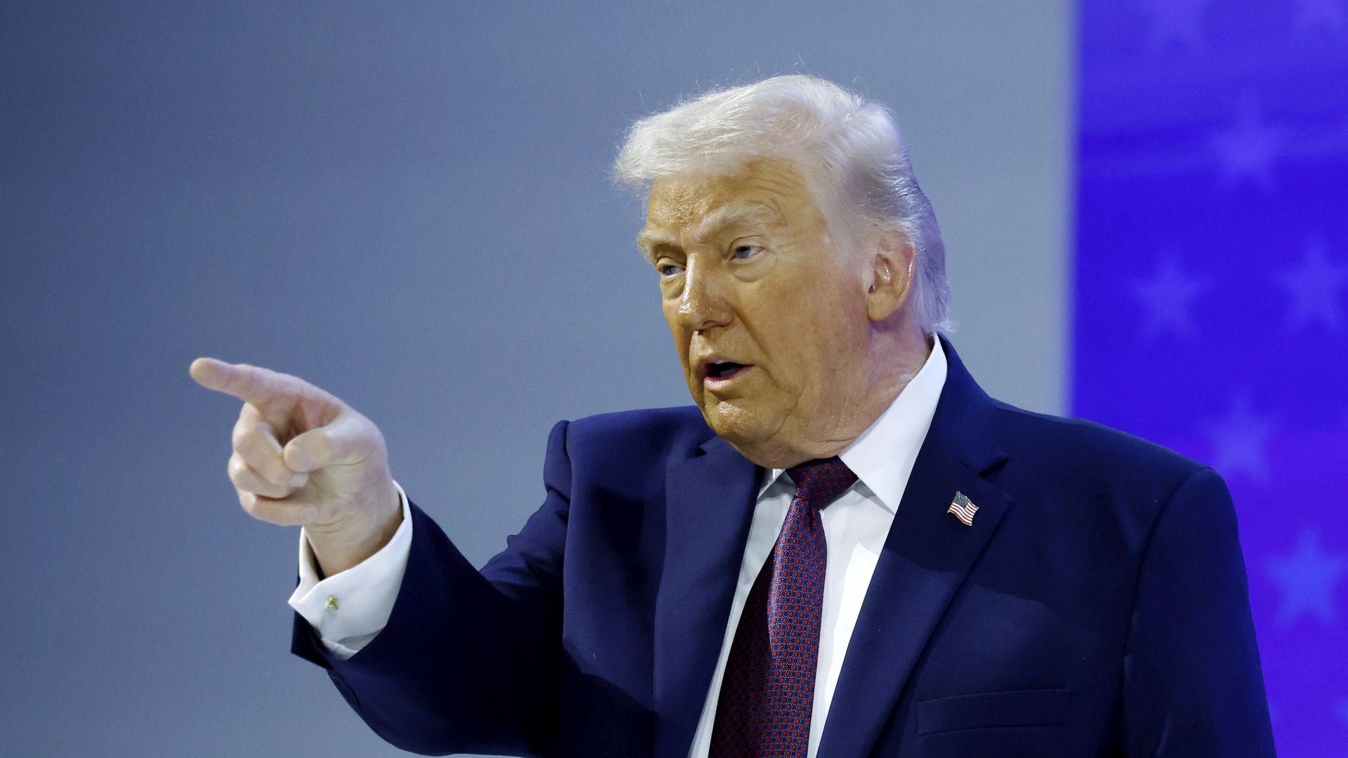 Trump pointing his finger into the crowd wearing a blue suit jacket with an American flag pin, white collared shirt and red and blue patterned tie
