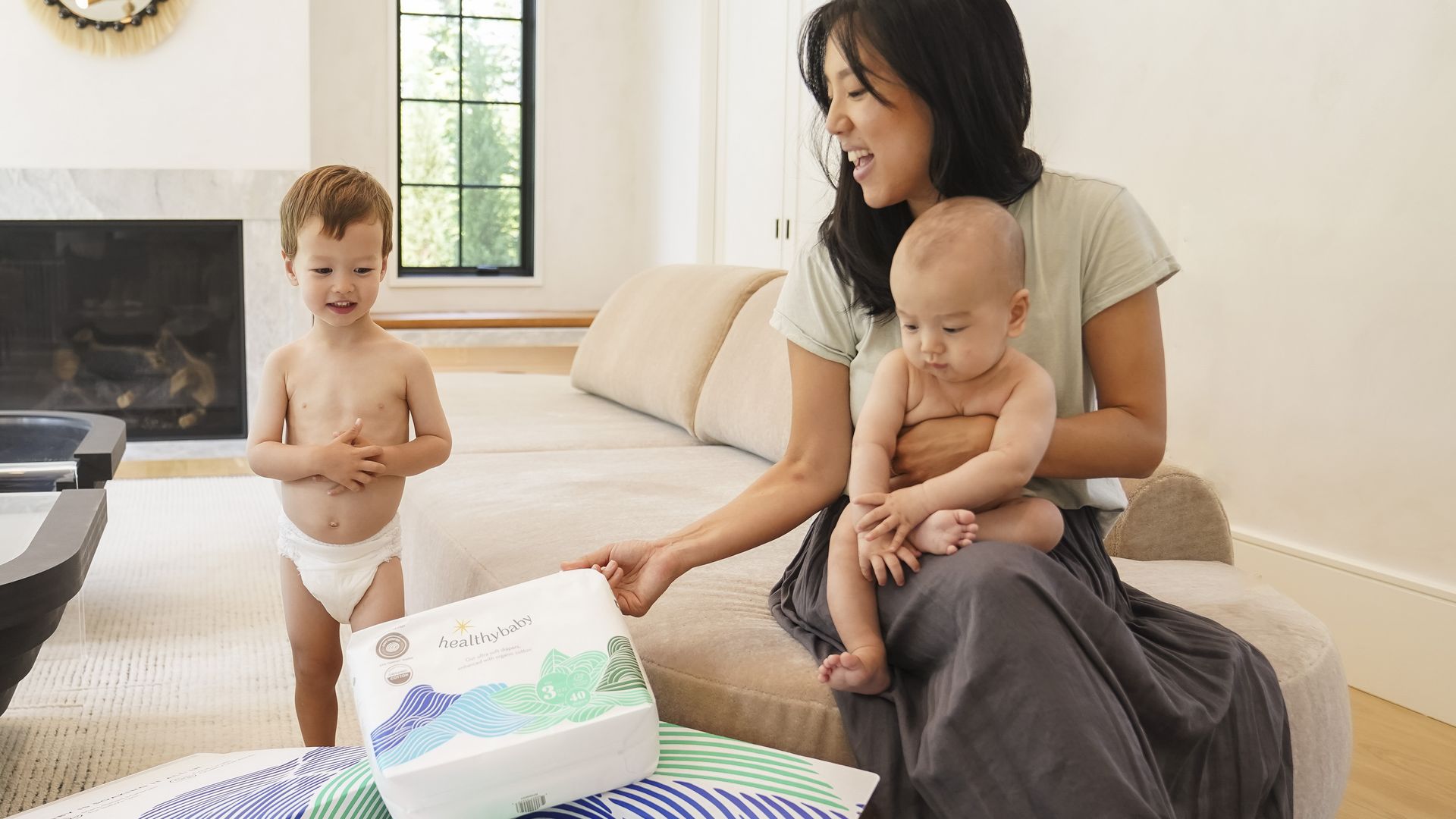 A mother with dark hair sits in a living room swathed in a blanket with her infant and toddler as she examines a package of Healthybaby diapers.