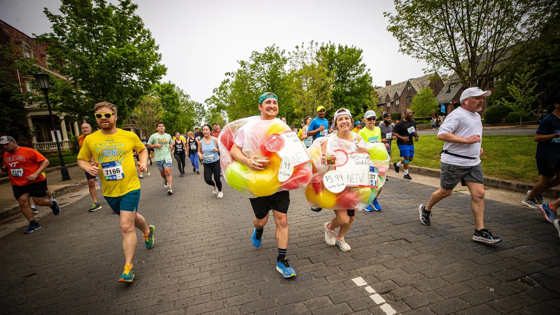 Runners race along a brick-paved street, lined with trees and houses. In the center, a man and woman wear colorful plastic balloon costumes, smiling as the crowd runs beside them.