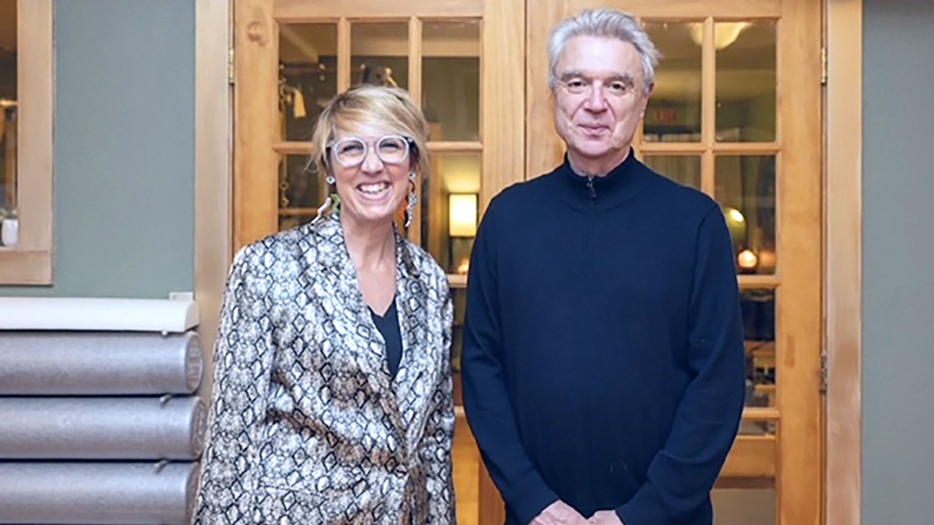 A woman in a black and white jecet and arty glasses poses with David Byrne in a navy nehru jacket