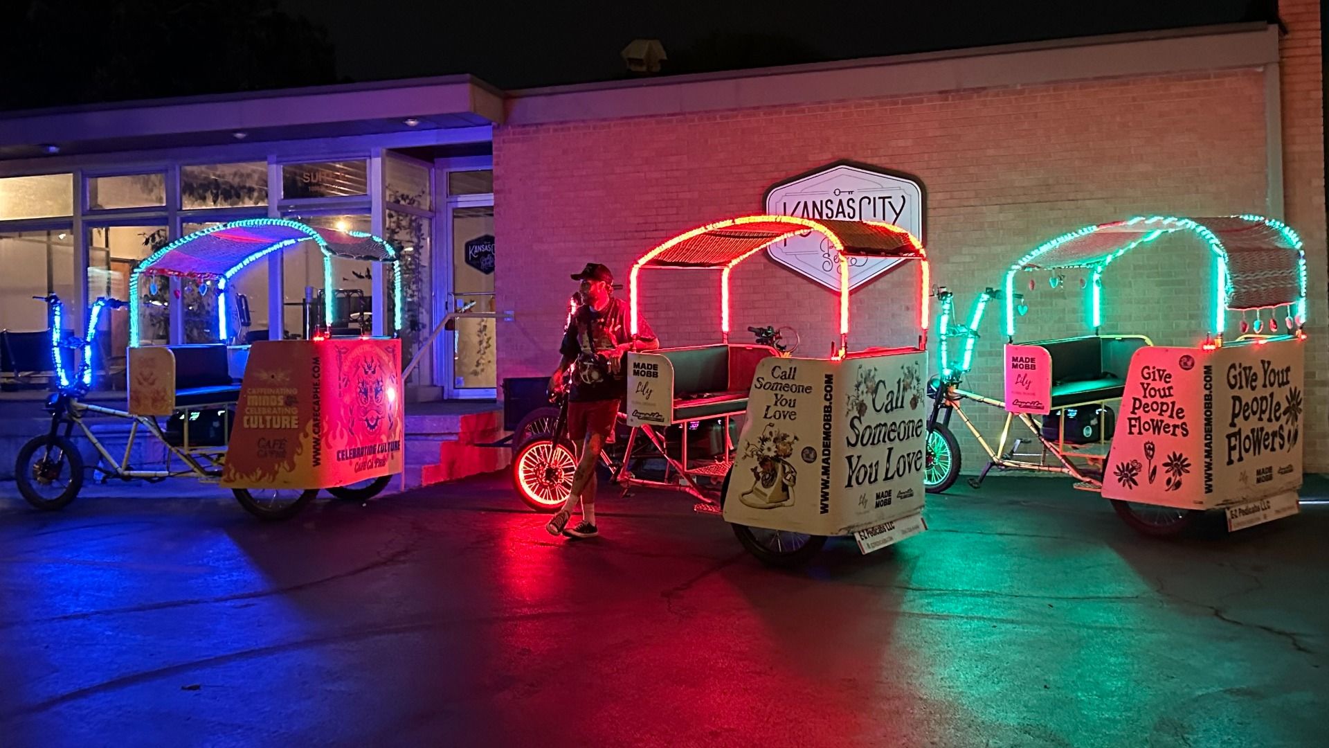 Three E-Z Pedicab vehicles lit up in blue, red, and green LED lights parked outside a building with a sign reading "Kansas City" at night with a person standing near the red-lit one.