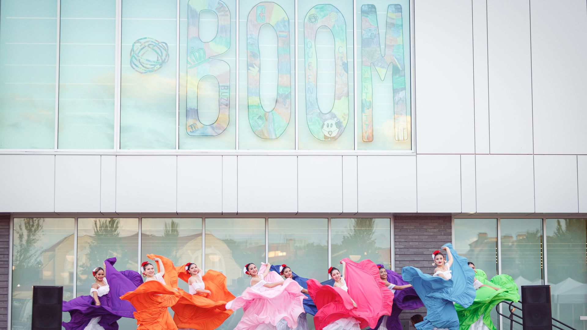 A group of dancers in bright, flowing dresses perform on an outdoor stage in front of a modern glass building; large multicolored letters spell "COLOR" across the windows.