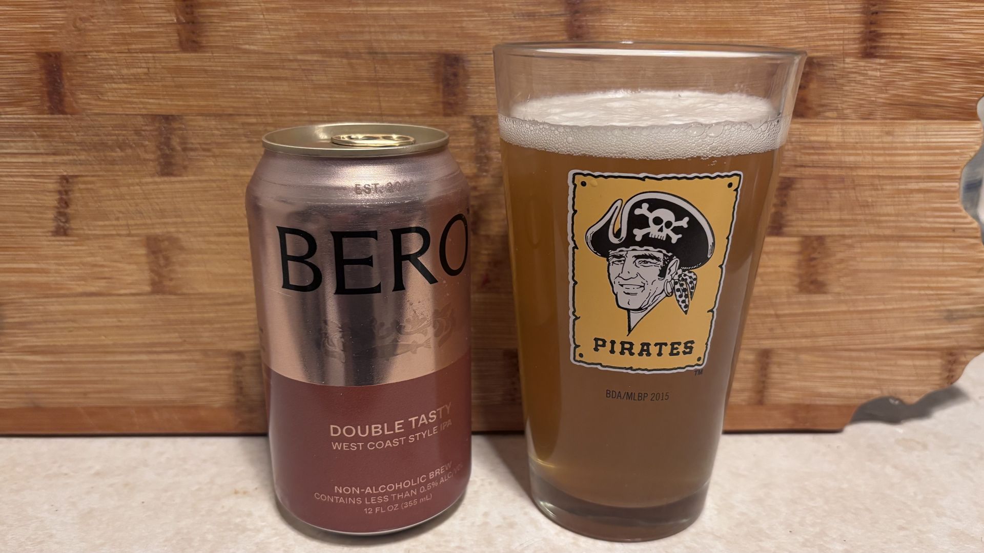 A can of Bero Double Tasty West Coast Style IPA non-alcoholic brew next to a glass filled with beer, featuring a Pittsburgh Pirates logo, on a beige countertop with wood background.