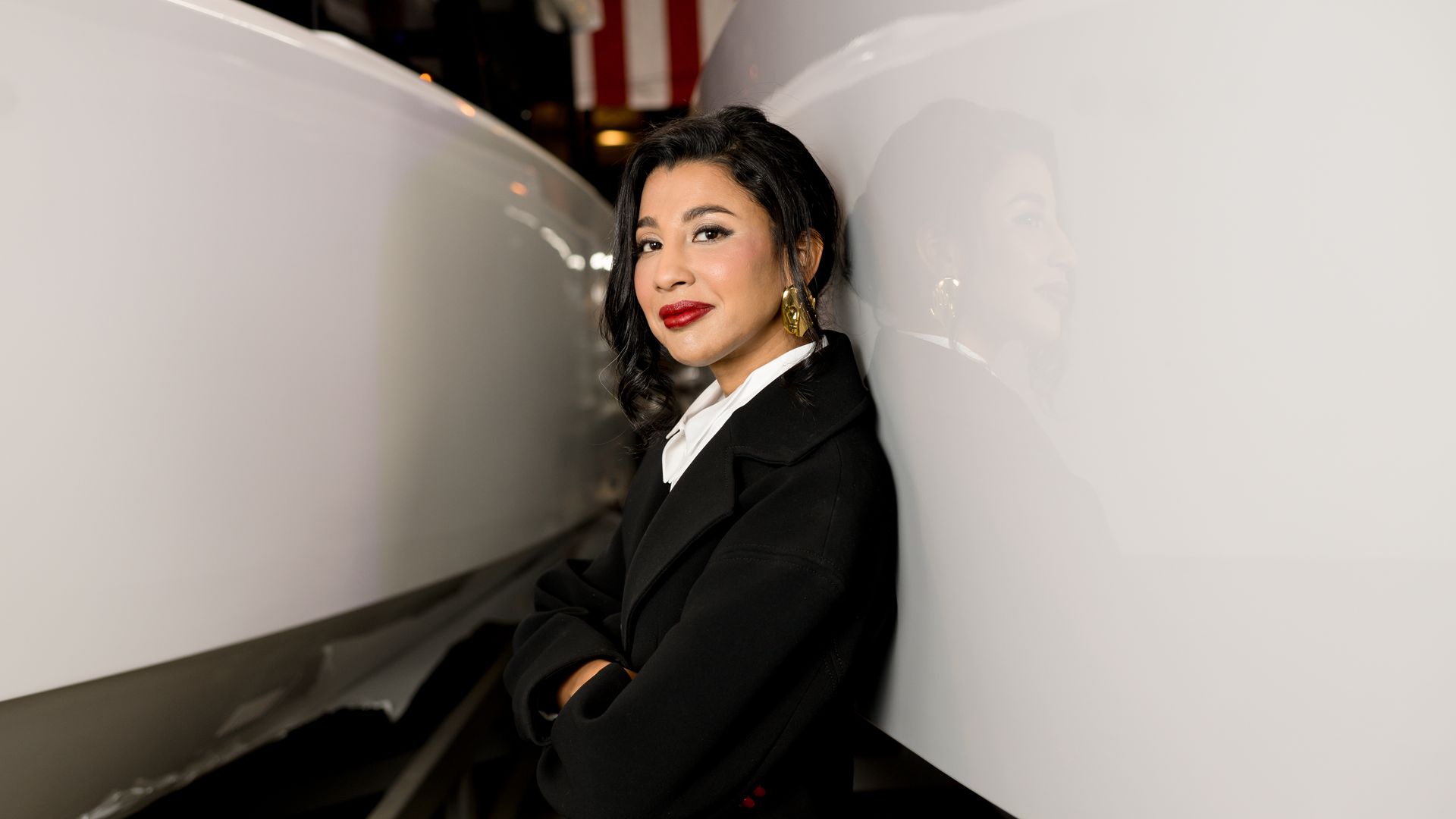 Woman with dark hair, red lipstick, and gold earrings, wearing a black coat and white shirt, stands between two large white surfaces, leaning against one with her arms crossed.