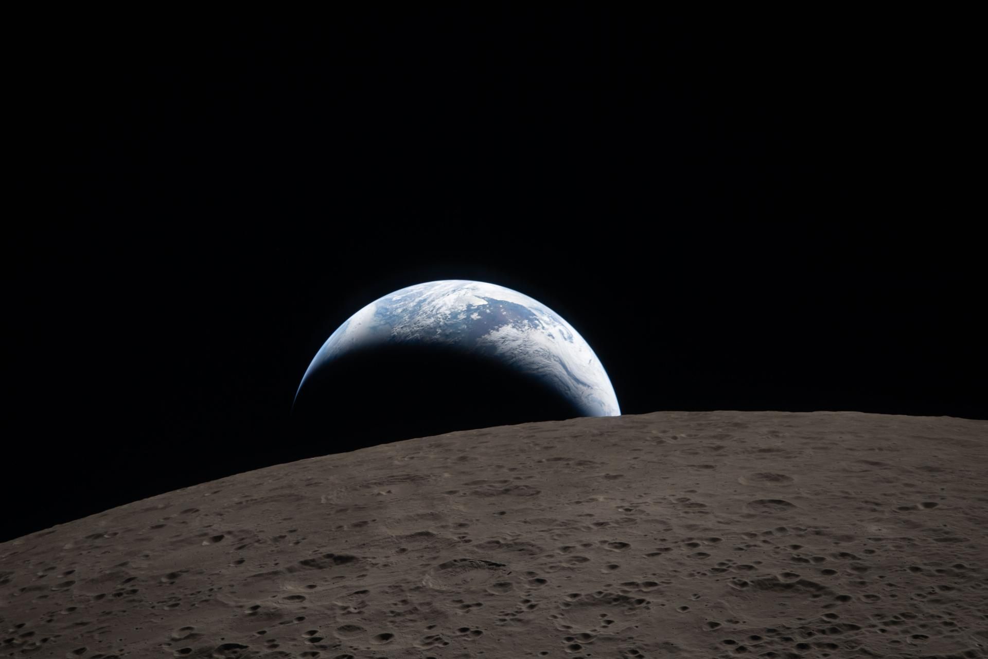 Earthset captured through the Orion spacecraft window at 6:41 p.m. EDT, April 6, 2026, during the Artemis II crew’s flyby of the Moon. A muted blue Earth with bright white clouds sets behind the cratered lunar surface.