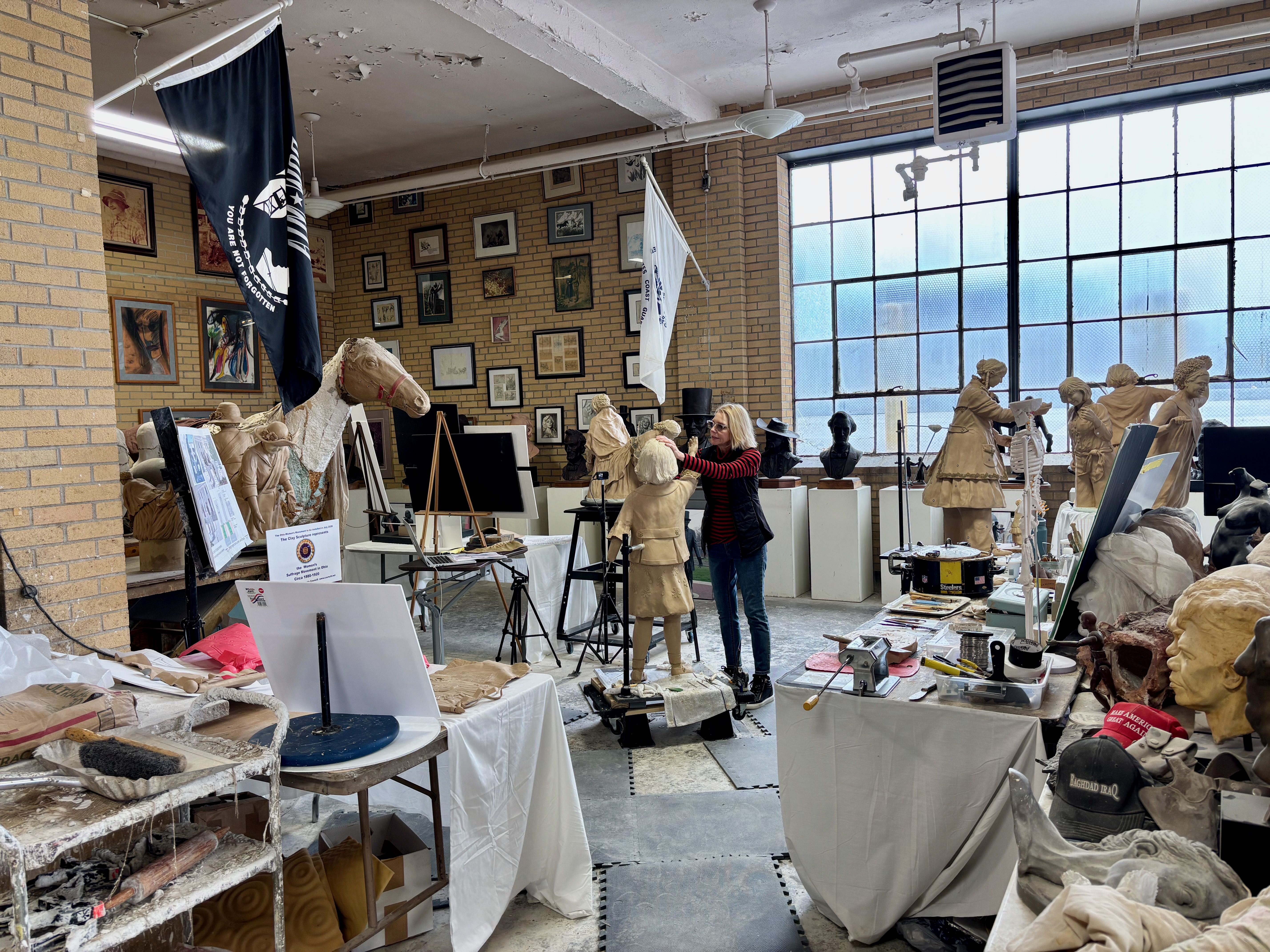 An art studio filled with plaster busts and statues; a woman adjusts a bust while others work. Brick walls, framed artworks, and large industrial windows with shelves and tables full of tools.