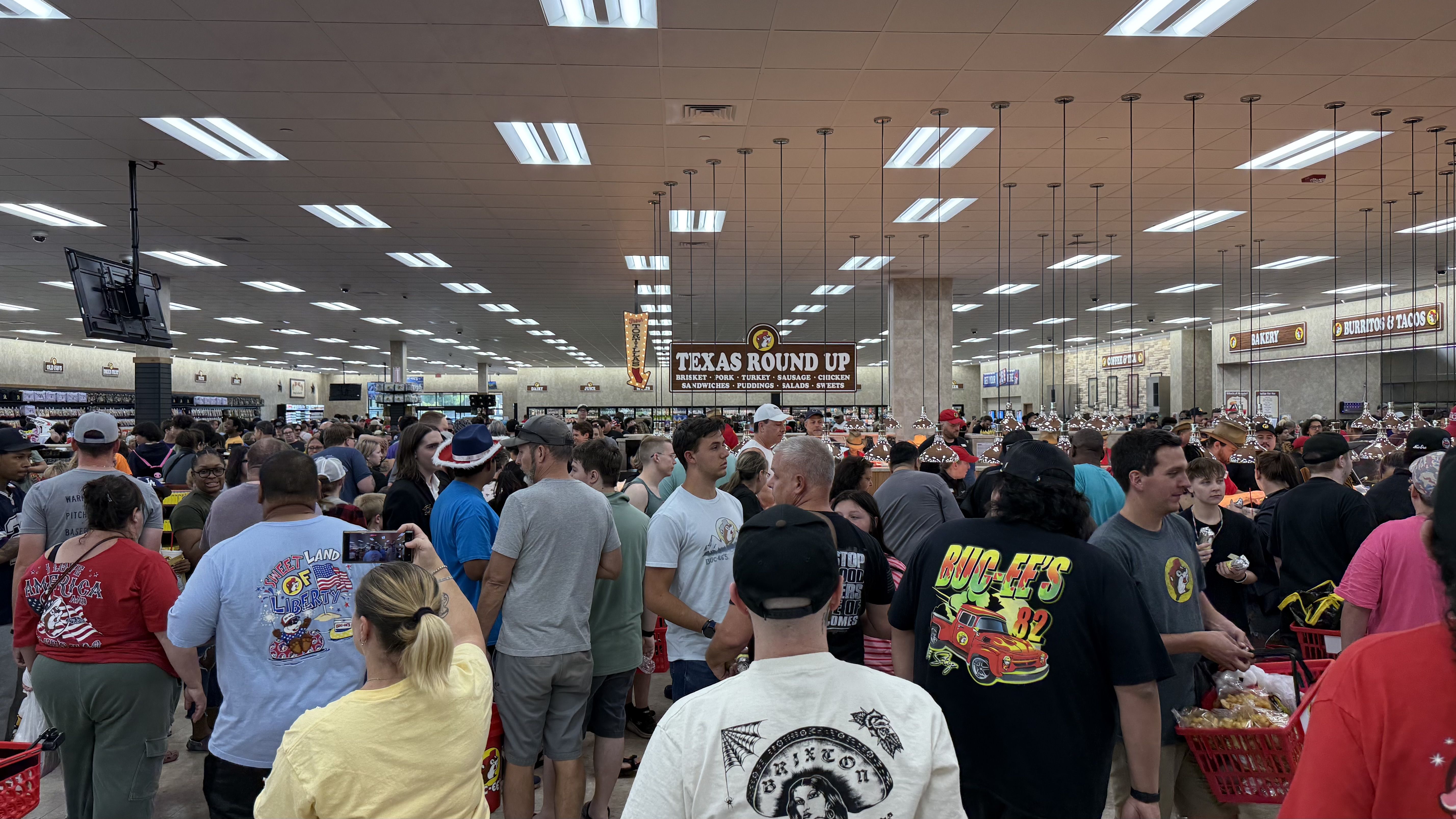 Hundreds of people walk around and shop inside Buc-ee's. A large sign reads "TEXAS ROUND UP" while bakery and burritos & tacos signs appear in the background.